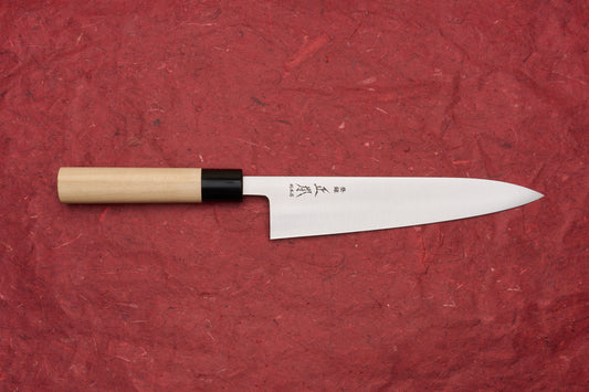 Masamoto KS Hongasumi Gyuto 210mm knife features a polished steel blade etched with Japanese characters and a light wood handle, showcasing its precision and craftsmanship on a red textured surface.