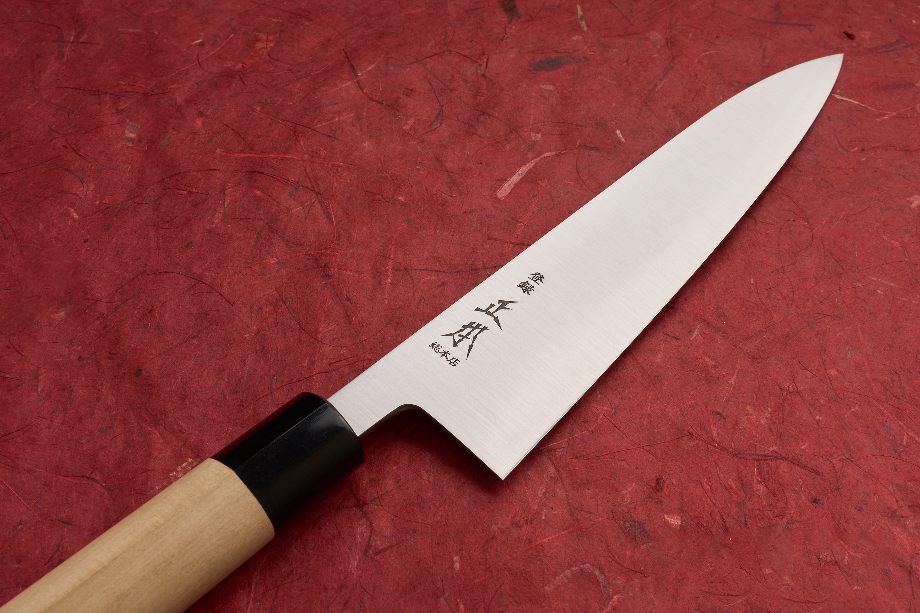 Masamoto KS Hongasumi Gyuto 210mm knife with a stainless-steel blade engraved with Japanese characters, resting on a deep red textured background, showcasing its sleek design and wooden handle with black bolster.