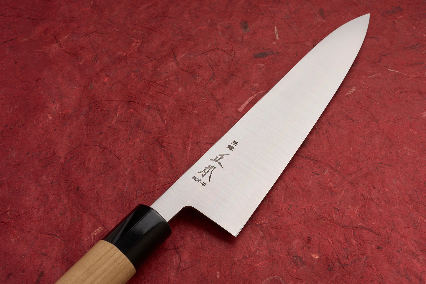 Masamoto KS Hongasumi Gyuto 240mm knife with a slender stainless-steel blade engraved with Japanese characters and an octagonal wooden handle, displayed on a red textured surface.
