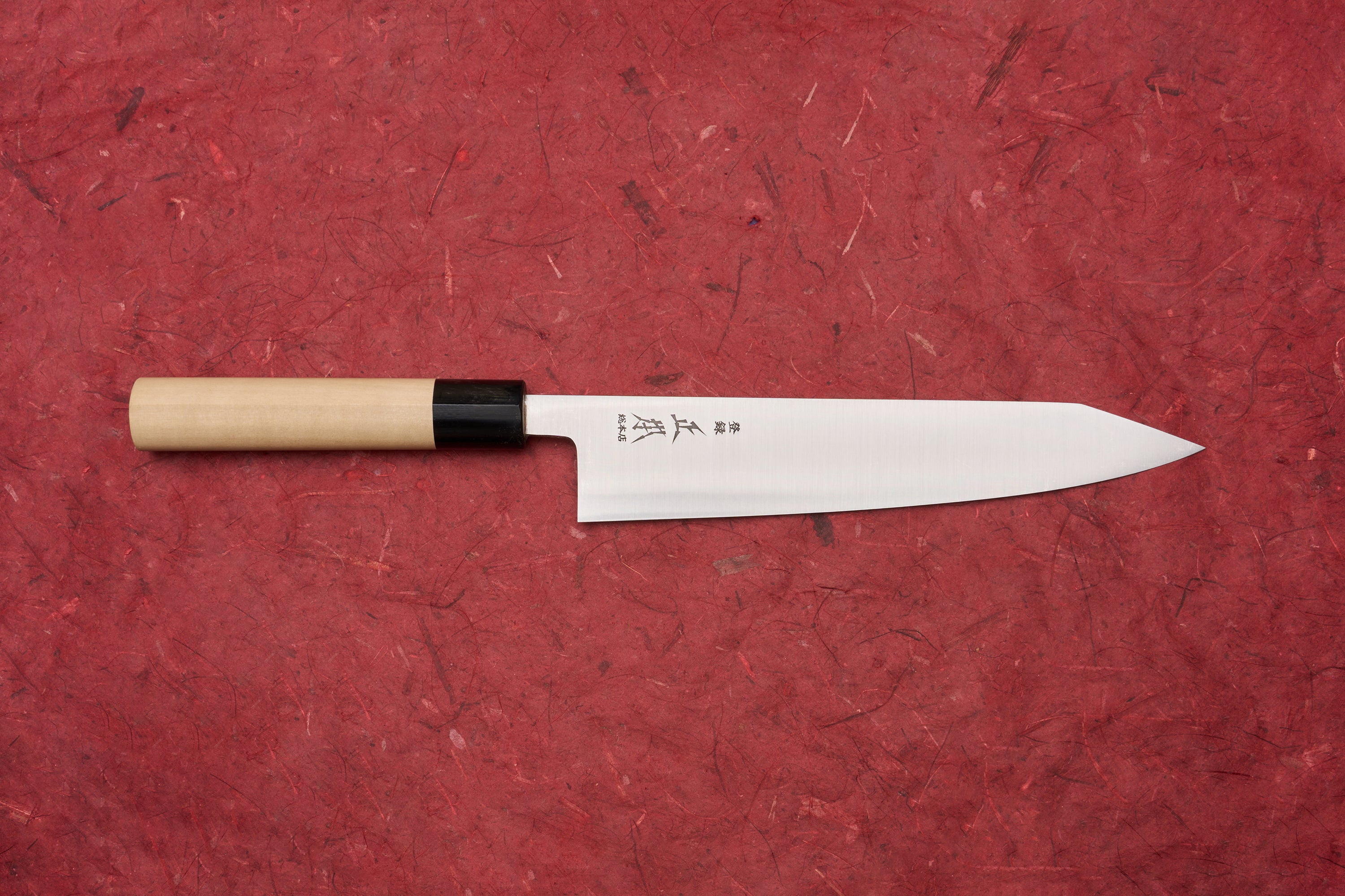 Masamoto KS Hongasumi Kiritsuke Gyuto 210mm knife with a polished, slender blade etched with Japanese characters and a wooden handle, displayed on a textured red background, embodying precision and craftsmanship.