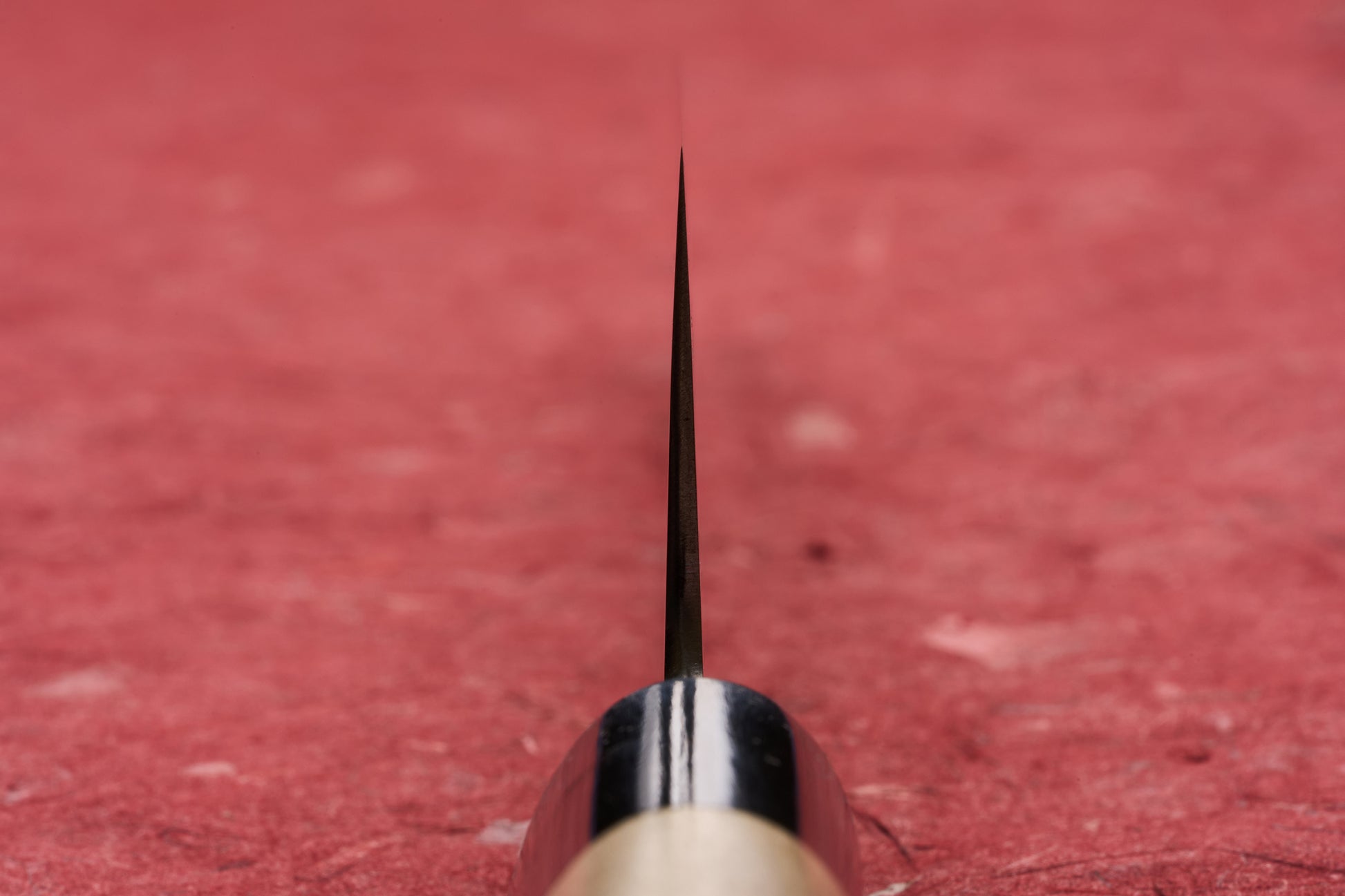 Close-up of the Masamoto KS Hongasumi Kiritsuke Gyuto 210mm, showcasing its sharp blade tip aimed at the camera, highlighting precision craftsmanship on a red textured surface.