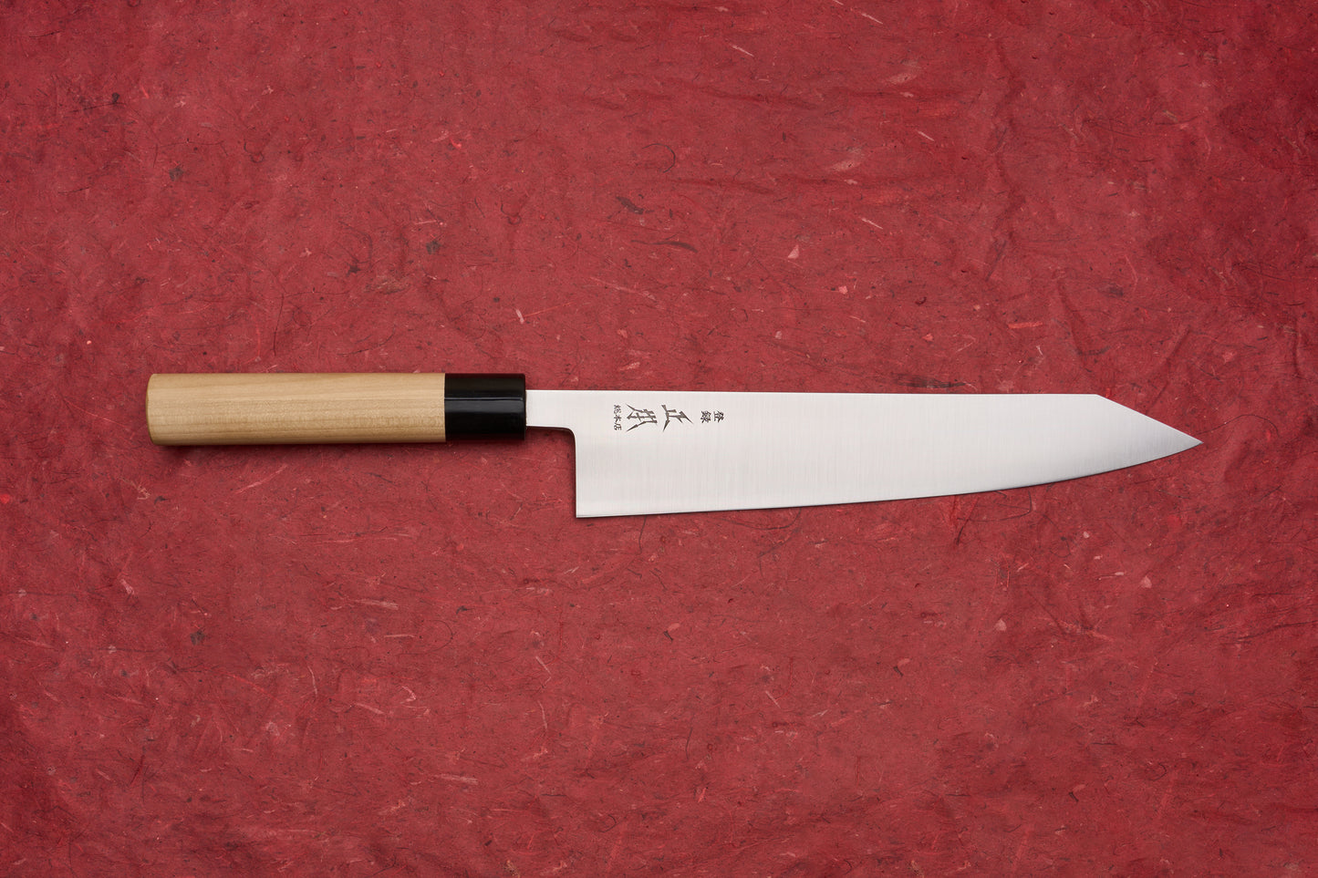 Masamoto KS Hongasumi Kiritsuke Gyuto 240mm knife with a polished, narrow blade featuring engraved Japanese characters, and a light wood handle, against a red textured background.