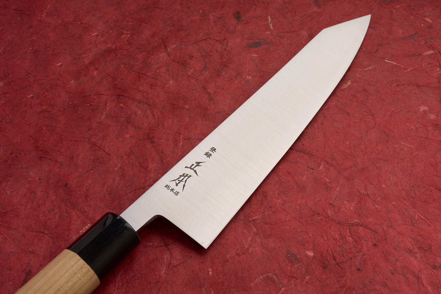 Masamoto KS Hongasumi Kiritsuke Gyuto 240mm knife with polished steel blade etched with Japanese characters, traditional wooden handle, and dark bolster, lying on a textured red surface.