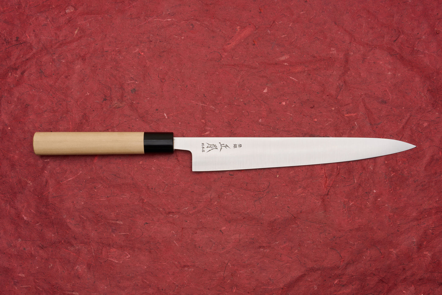 Masamoto KS Hongasumi Sujihiki 240mm knife with a sleek, single-bevel stainless blade and etched Japanese characters, featuring a light wood handle and black ferrule, displayed on a red textured background.