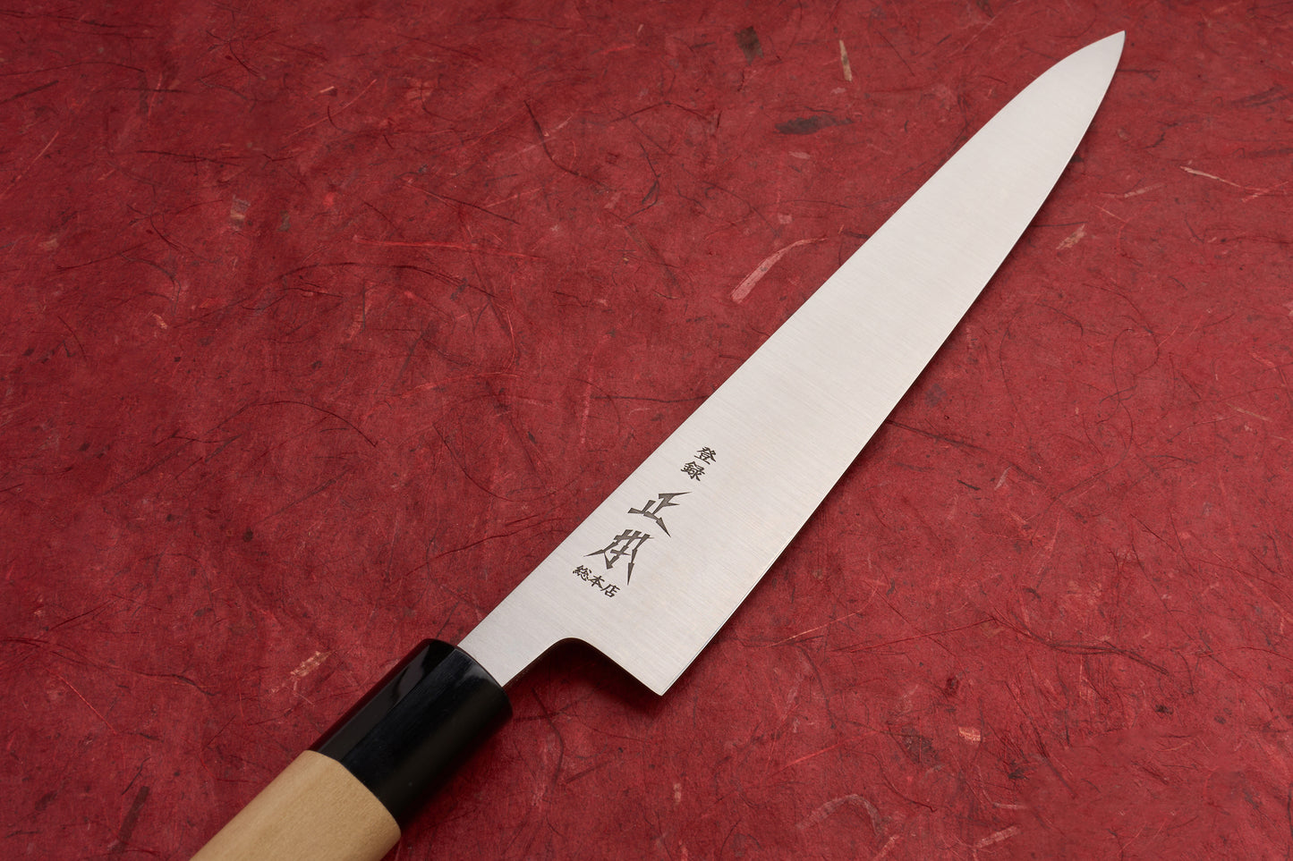 Masamoto KS Hongasumi Sujihiki 240mm knife features a polished stainless steel yanagiba blade with Japanese kanji, paired with an octagonal wooden handle and black ferrule, on a red fibrous paper background.