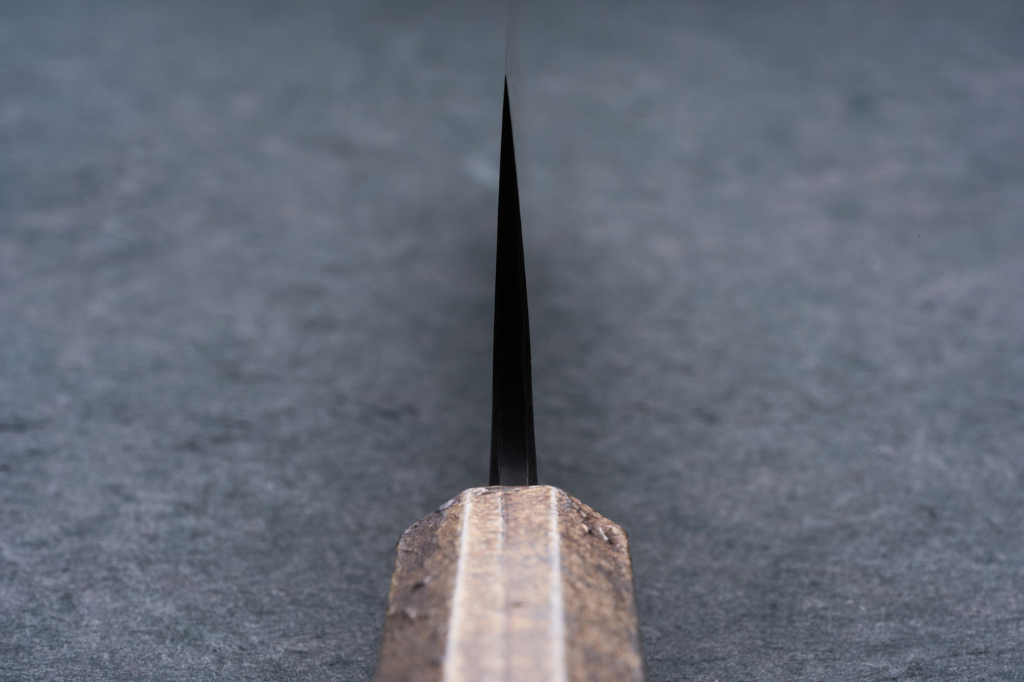 Mazaki Aoichi Beads Black Damascus Gyuto 240mm knife blade emerging from a wooden handle, set on a gray surface, showcasing Japanese craftsmanship.
