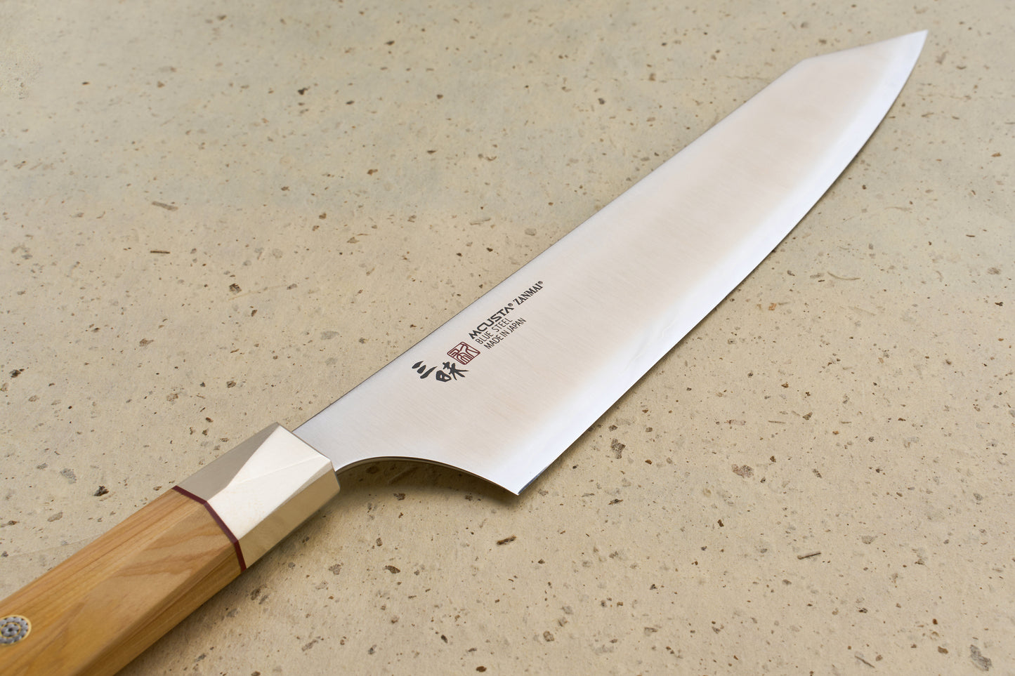 Mcusta Zanmai Beyond Kiritsuke Gyuto 210mm knife featuring a polished carbon-steel blade with Japanese etchings and an octagonal wooden handle, displayed on a light speckled countertop.