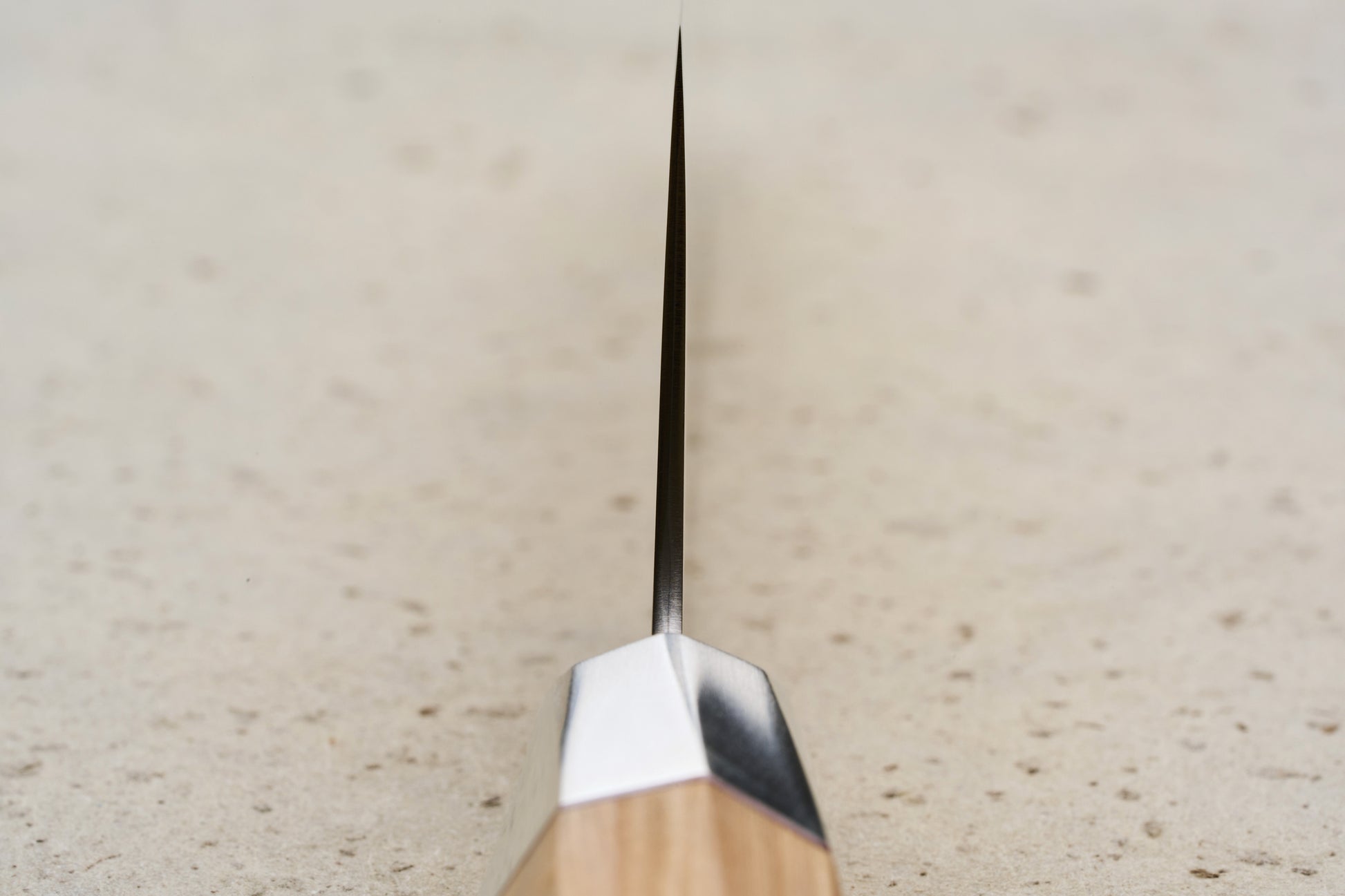 Mcusta Zanmai Beyond Kiritsuke Gyuto 210mm knife, showcasing its thin steel blade, metal bolster, and wooden handle, set edge-on against a light speckled surface, embodying precision craftsmanship.