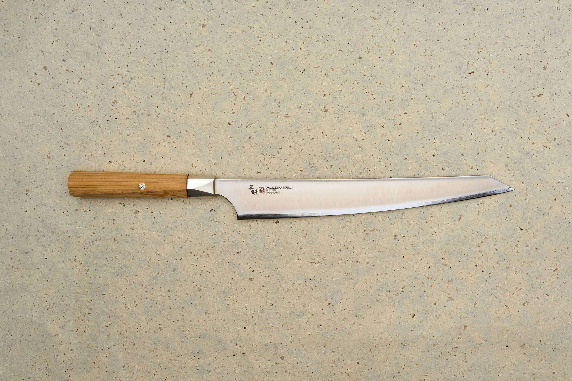 Mcusta Zanmai Beyond Kiritsuke Sujihiki 270mm knife on a stone surface, showcasing its high-carbon steel blade with hamon and octagonal wooden handle, featuring detailed maker's inscription and Japanese characters.