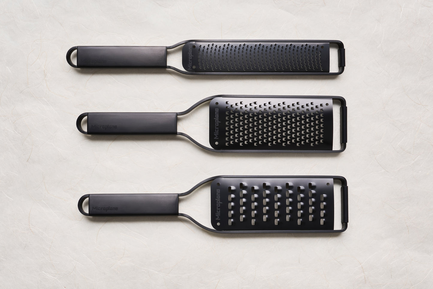 Overhead view of Microplane Black Sheep Series Graters: fine rasp/zester, medium-hole, and coarse-hole graters, each with a long grating surface and slim handle with hanging loop.