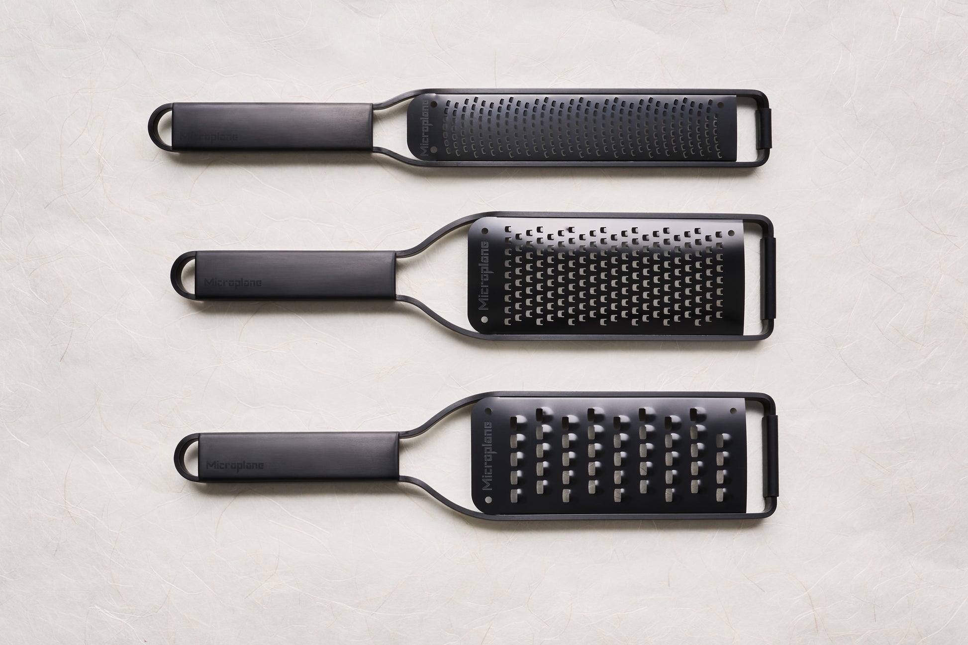 Overhead view of Microplane Black Sheep Series Graters: fine rasp/zester, medium-hole, and coarse-hole graters, each with a long grating surface and slim handle with hanging loop.