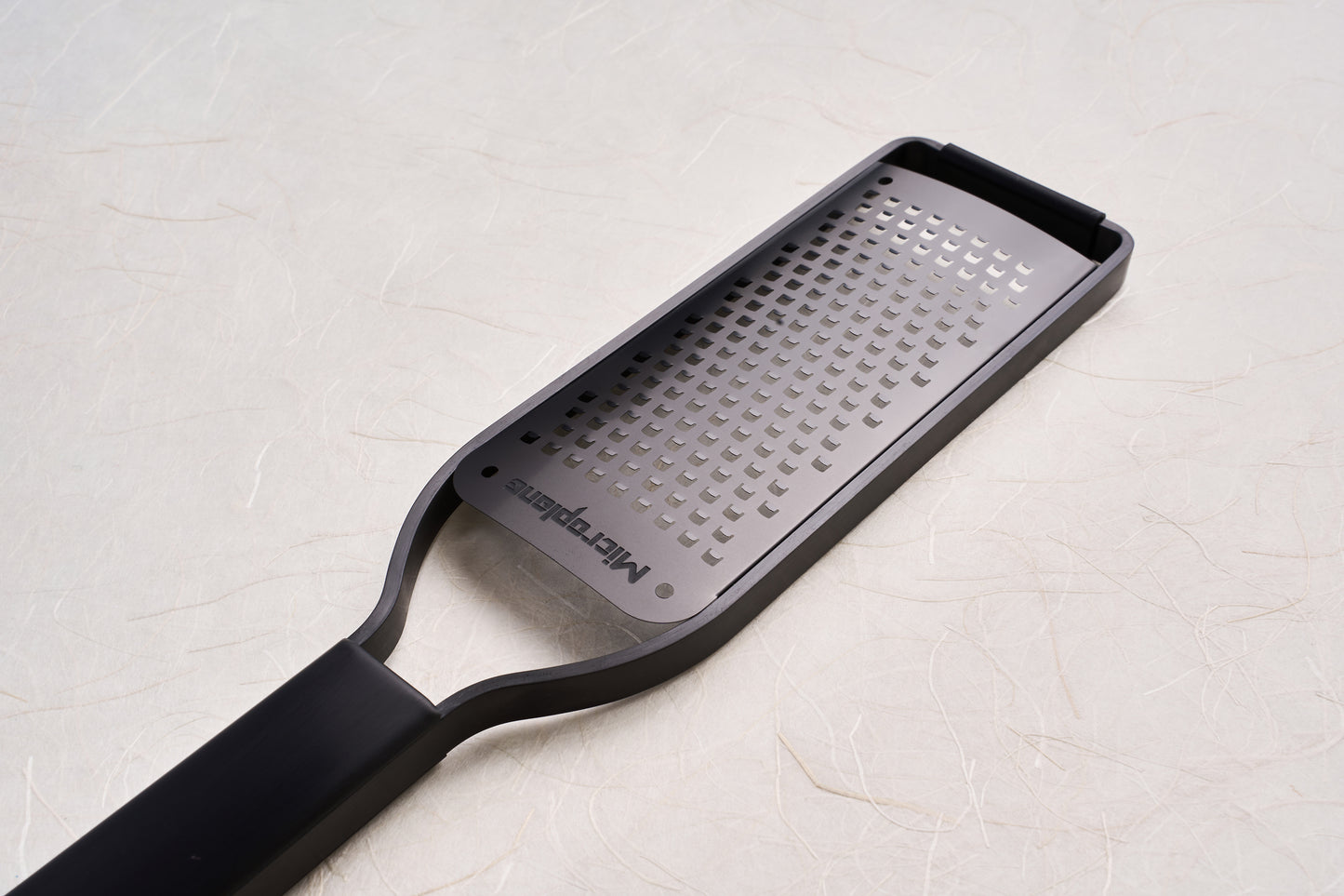 Microplane Black Sheep Series Grater with a perforated stainless-steel blade and black plastic handle, ideal for grating hard cheeses, spices, and citrus, showcased on a textured surface.