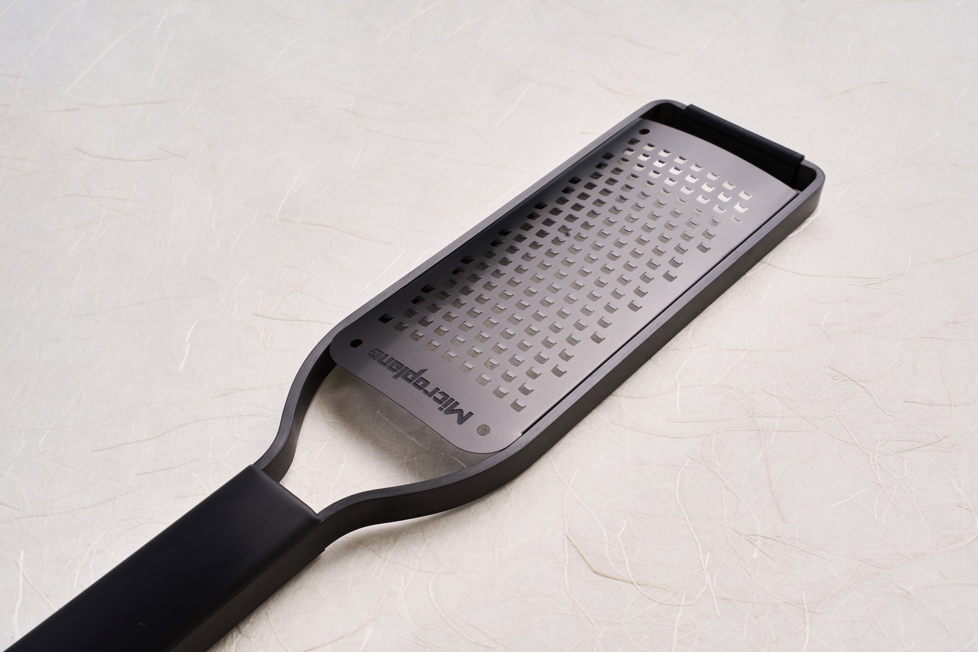 Microplane Black Sheep Series Grater with a perforated stainless-steel blade and black plastic handle, ideal for grating hard cheeses, spices, and citrus, showcased on a textured surface.