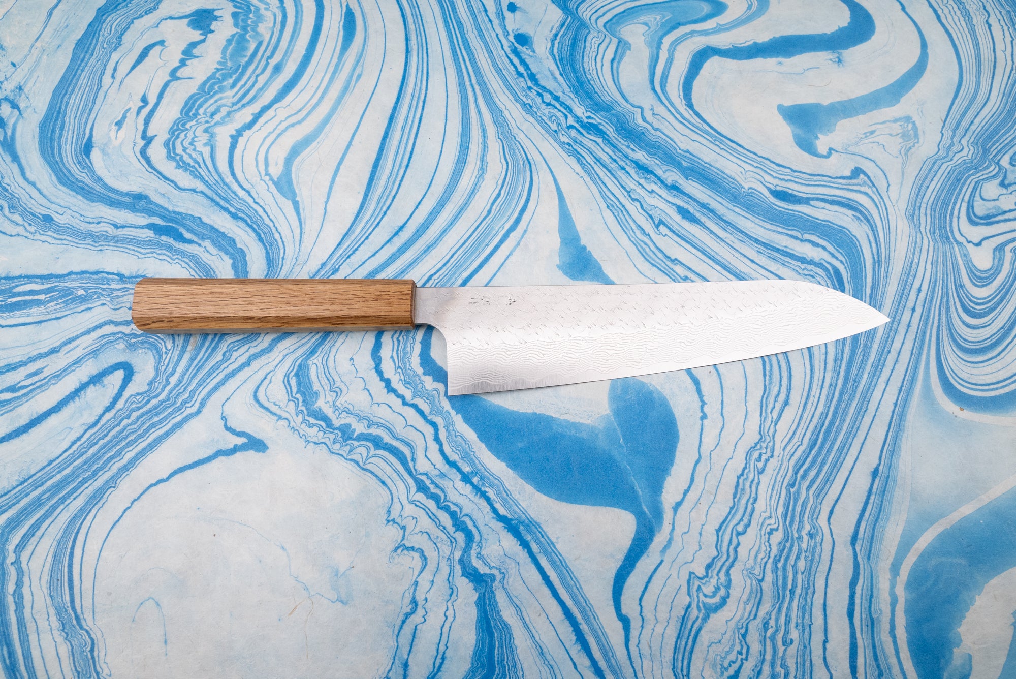 Nigara SPG STRIX Tsuchime Damascus Gyuto 240mm, showcasing a beautifully crafted Japanese-style blade with a subtle Damascus pattern, laid flat with a straight wooden handle on a marbled surface.