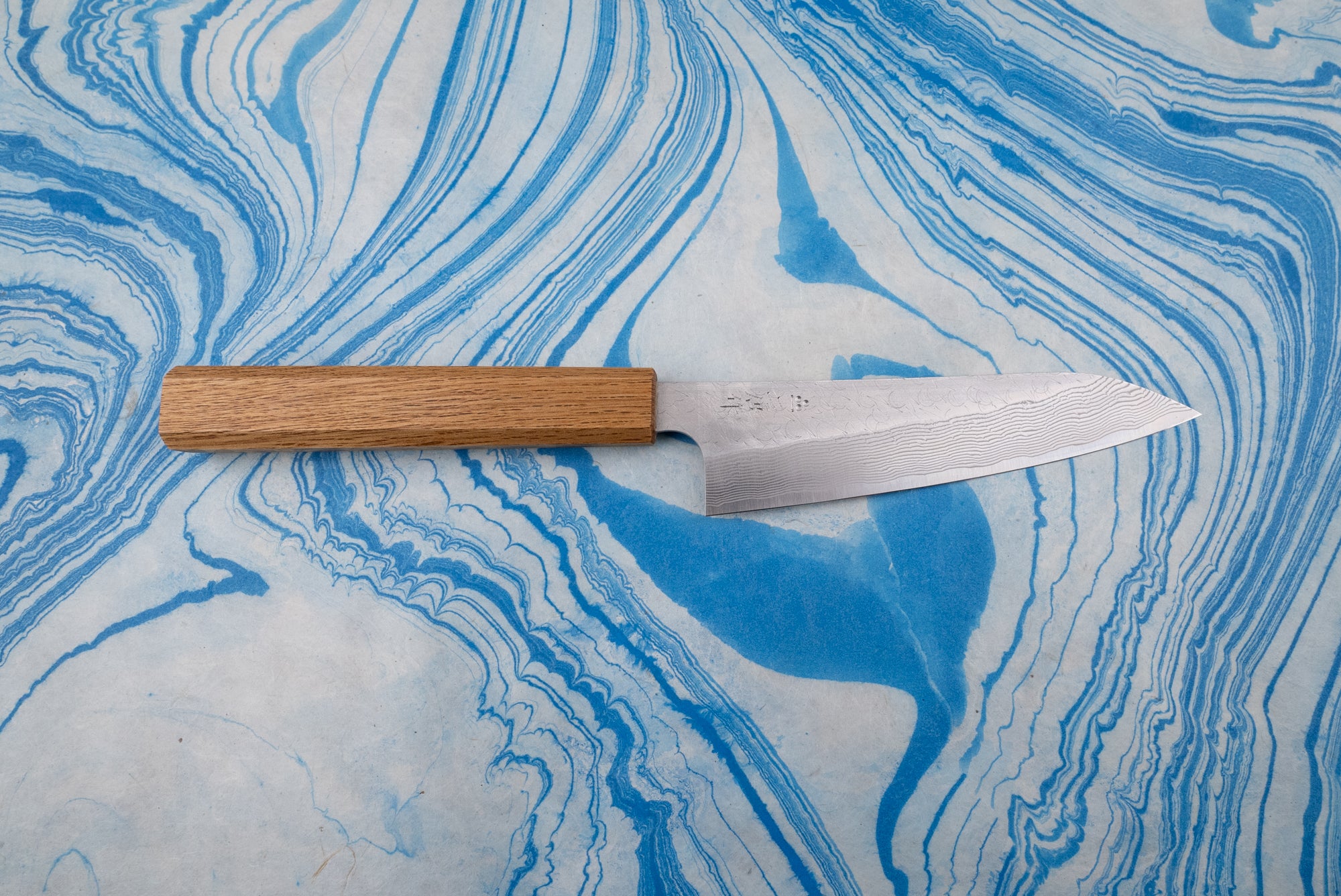 Nigara SPG STRIX Tsuchime Damascus Petty 150mm knife with a narrow blade featuring a subtle Damascus pattern and etched characters, paired with an octagonal wooden handle, lying on a marbled surface.