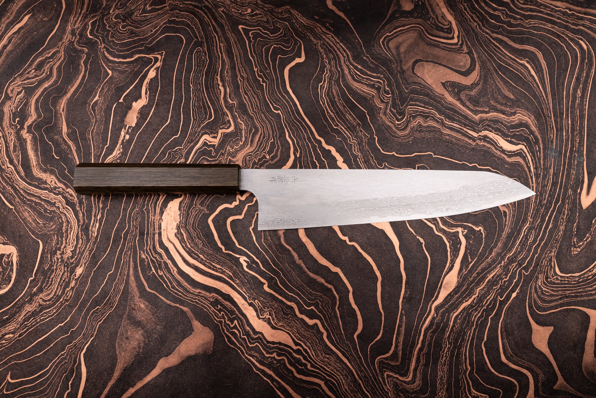 Nigara VG XEOS Tsuchime Damascus Gyuto 255mm with a damascus-patterned blade and dark wooden octagonal handle on a marbled, copper-accented surface, exemplifying Japanese craftsmanship from Aomori's historic knifemaker.