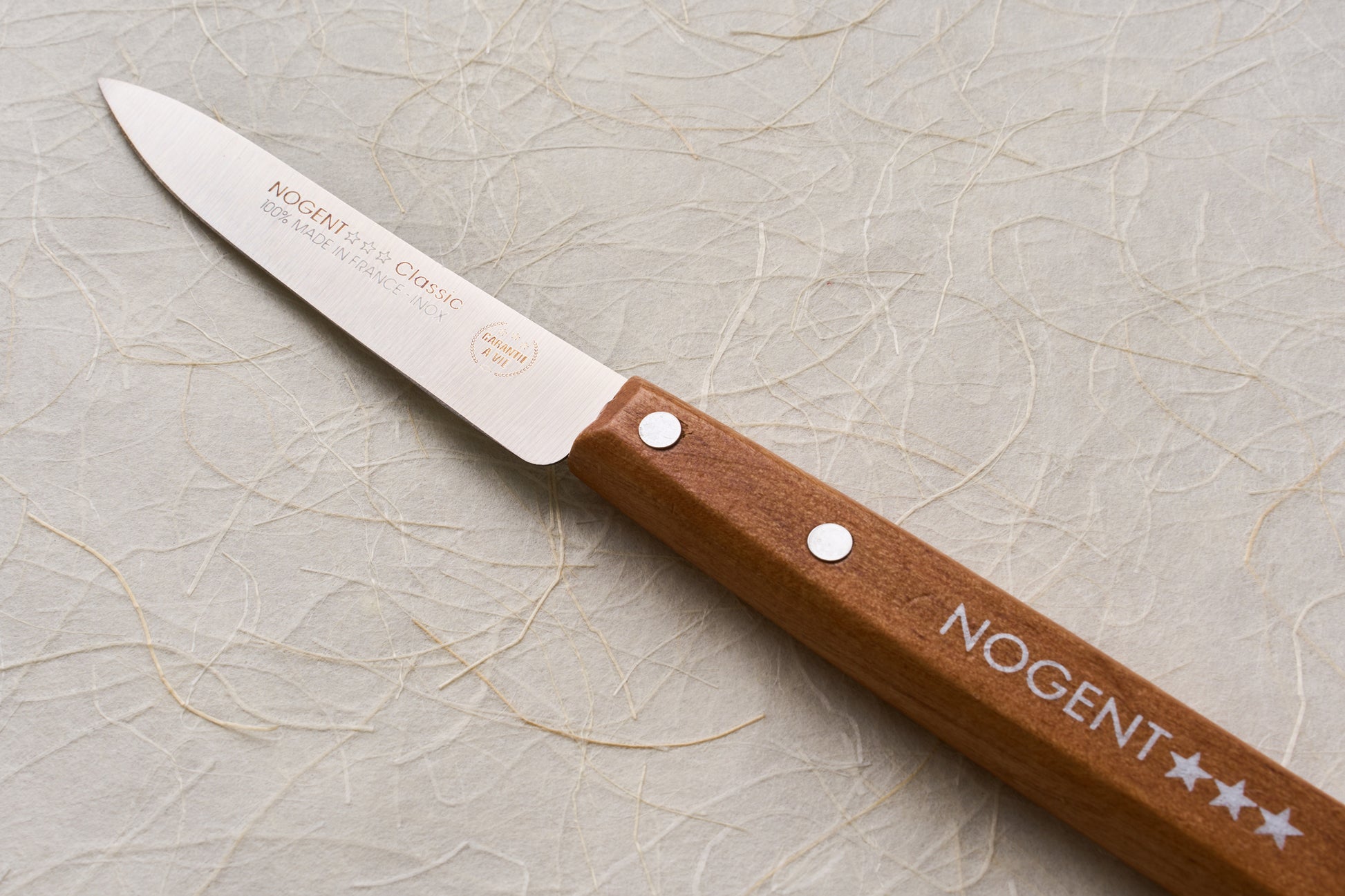 Nogent Paring Knife with a stainless steel blade and branded wooden handle, embodying French craftsmanship and tradition, displayed on a textured paper background.
