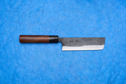 Onihiro Shiroichi Kurouchi Nakiri 135mm, a Japanese-style vegetable knife with a rectangular dual-tone blade and wooden handle, ideal for precise push/pull chopping, displayed on a blue background.