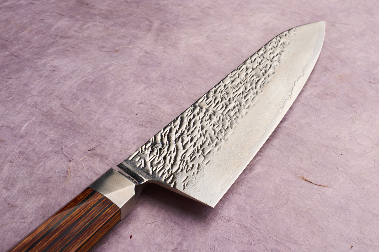 Ono Knife Suzukaze SG2 Tsuchime Damascus Gyuto 210mm with hammered blade texture and Damascus layering, polished steel bolster, and straight-grained wooden handle, displayed on a textured lavender surface.