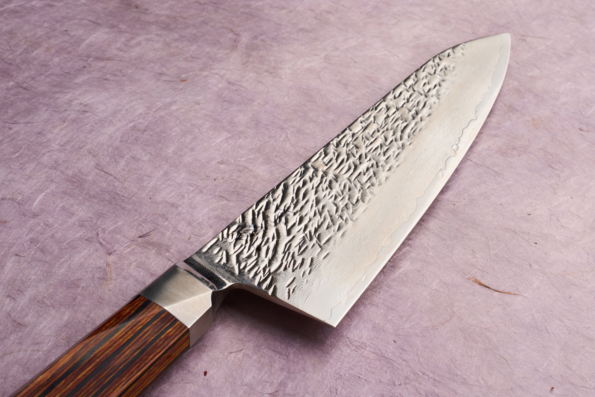 Ono Knife Suzukaze SG2 Tsuchime Damascus Gyuto 210mm with hammered blade texture and Damascus layering, polished steel bolster, and straight-grained wooden handle, displayed on a textured lavender surface.