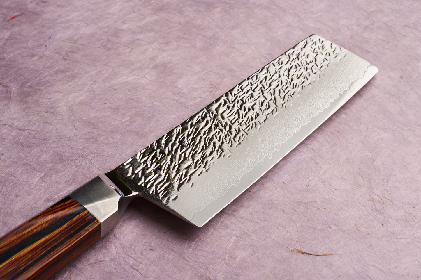 Ono Knife Suzukaze SG2 Tsuchime Damascus Nakiri 165mm, featuring a hammered, Damascus-patterned steel blade with a polished bolster and a striped wooden handle, lies on a textured surface.