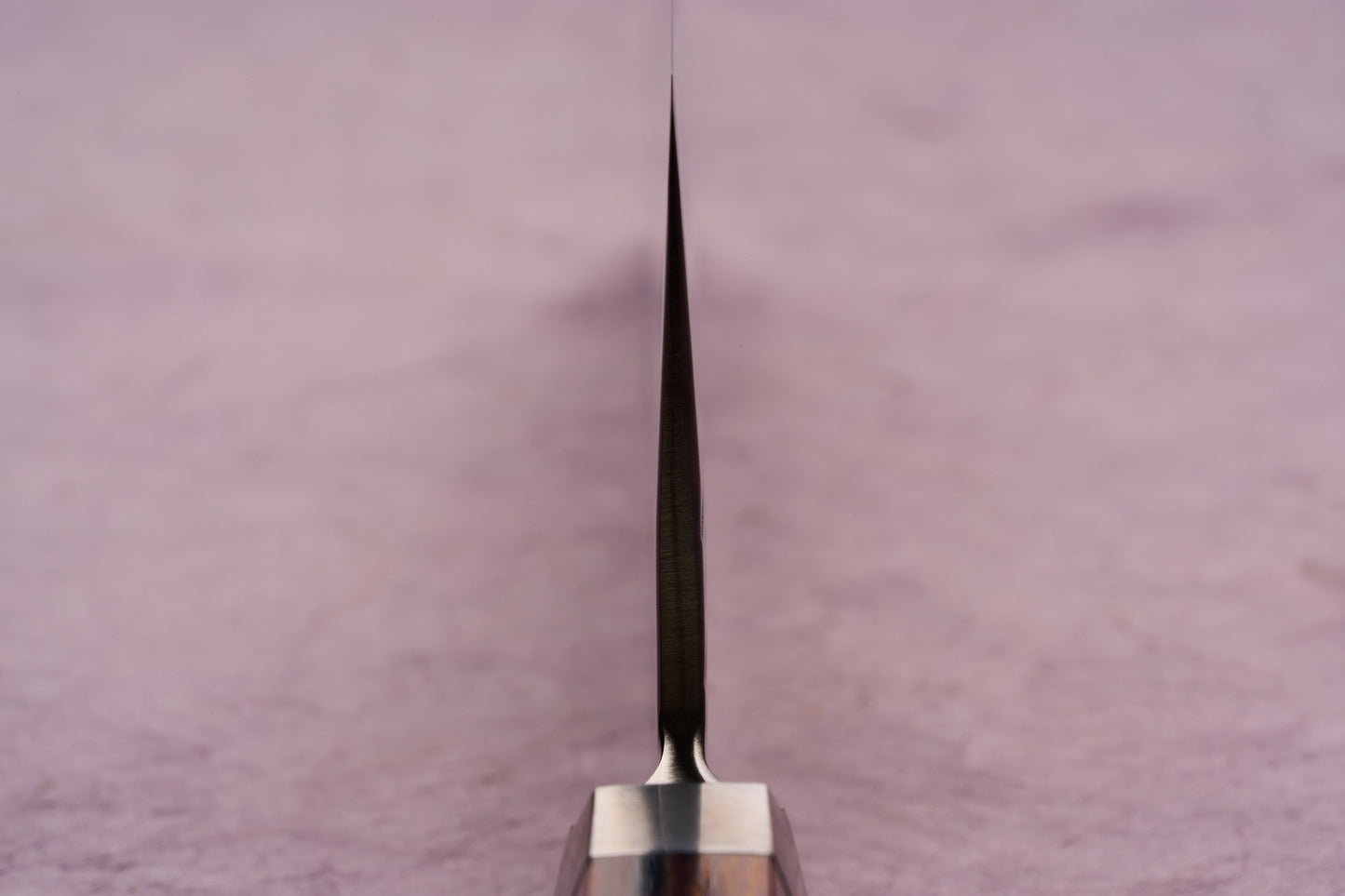 The Ono Knife Suzukaze SG2 Tsuchime Damascus Nakiri 165mm is shown with its sharp blade tip and spine in focus, highlighting its precision craftsmanship and sleek design on a pale pink surface.