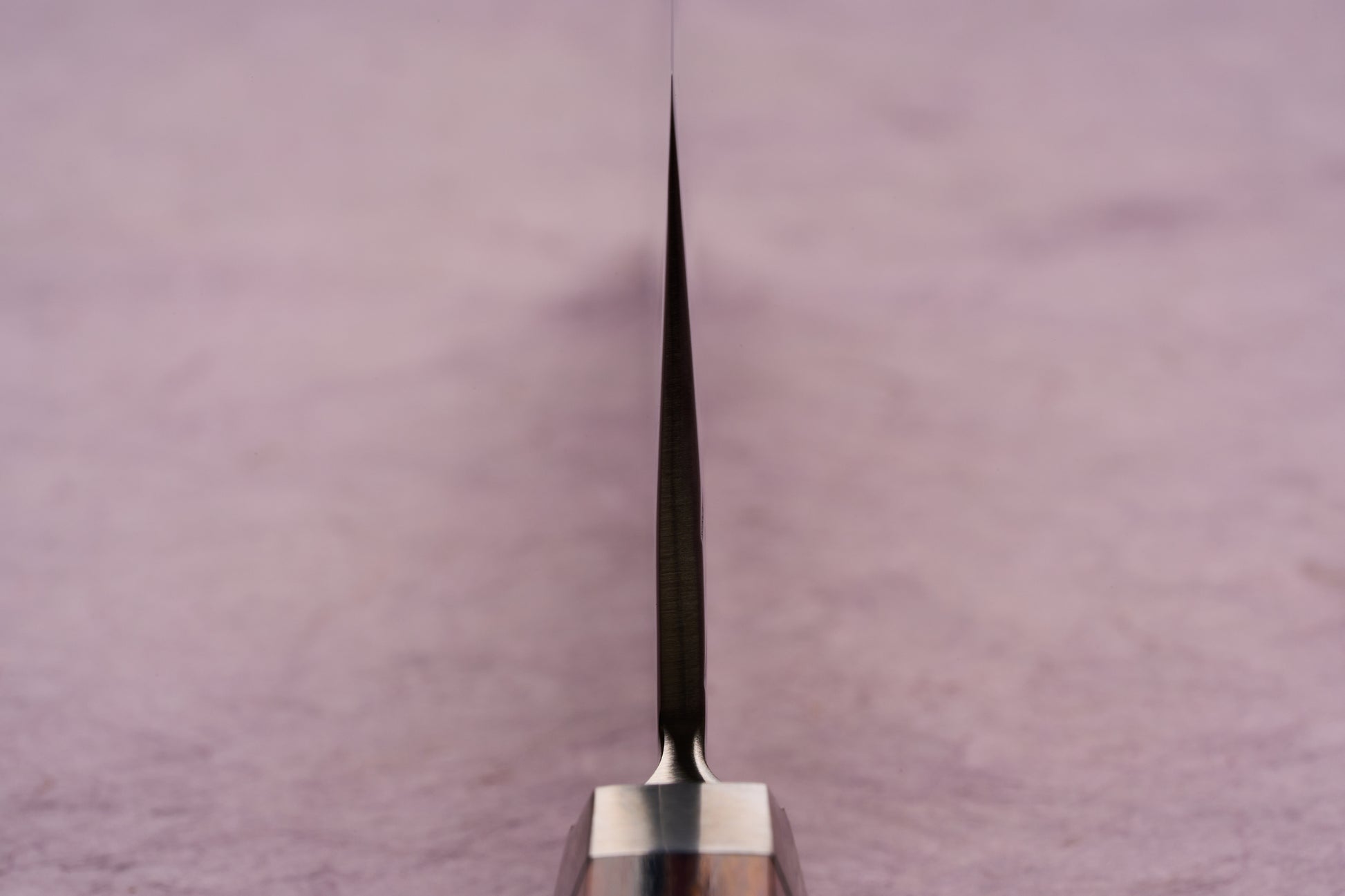 The Ono Knife Suzukaze SG2 Tsuchime Damascus Nakiri 165mm is shown with its sharp blade tip and spine in focus, highlighting its precision craftsmanship and sleek design on a pale pink surface.