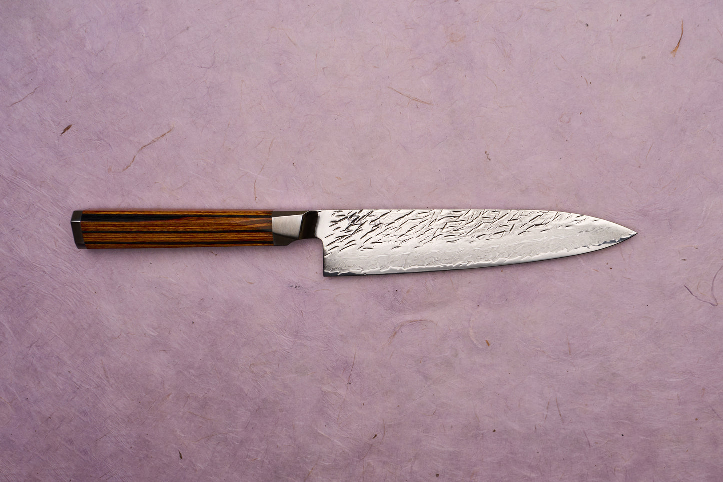 Ono Knife Suzukaze SG2 Tsuchime Damascus Petty 150mm showcases a hammered-finish Damascus blade with a striped wood handle, embodying precision craftsmanship on a pale lavender-pink textured background.