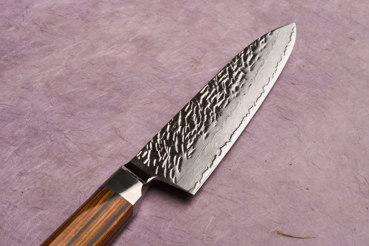 Ono Knife Suzukaze SG2 Tsuchime Damascus Petty 150mm, featuring a hammered steel blade with a wood-striped handle, exemplifies precision craftsmanship, ideal for delicate slicing tasks on a cutting board.