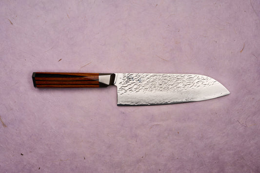 Ono Knife Suzukaze SG2 Tsuchime Damascus Santoku 180mm displayed on a textured surface, showcasing its hammered Damascus-style blade and dark octagonal wooden handle, embodying precision craftsmanship from Knifewear.