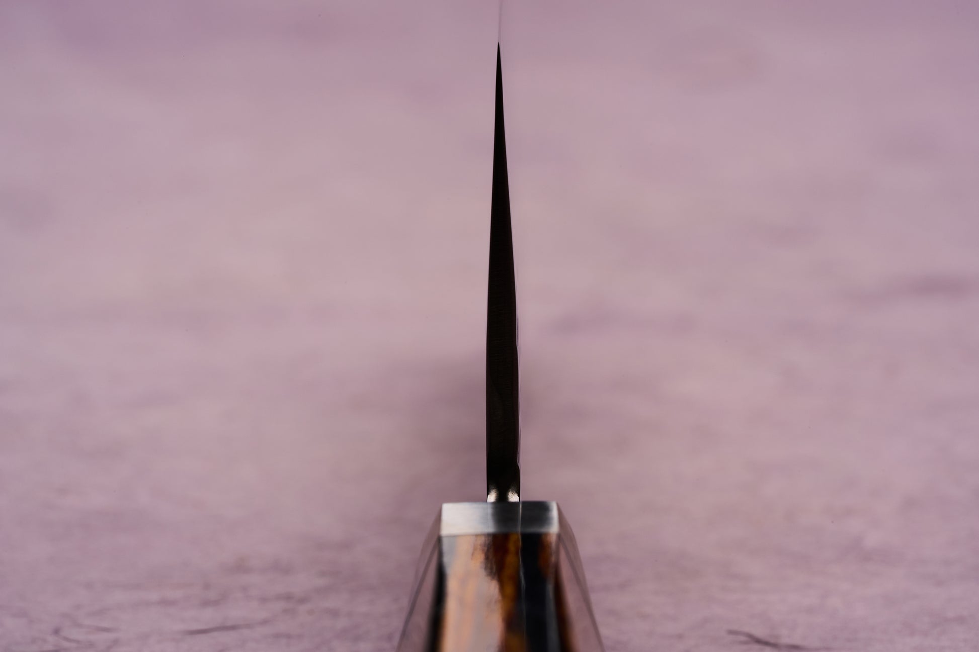 Ono Knife Suzukaze SG2 Tsuchime Damascus Santoku 180mm with a thin blade edge, wooden handle visible below, displayed against a soft pink background, showcasing precision craftsmanship and cutting-edge design.