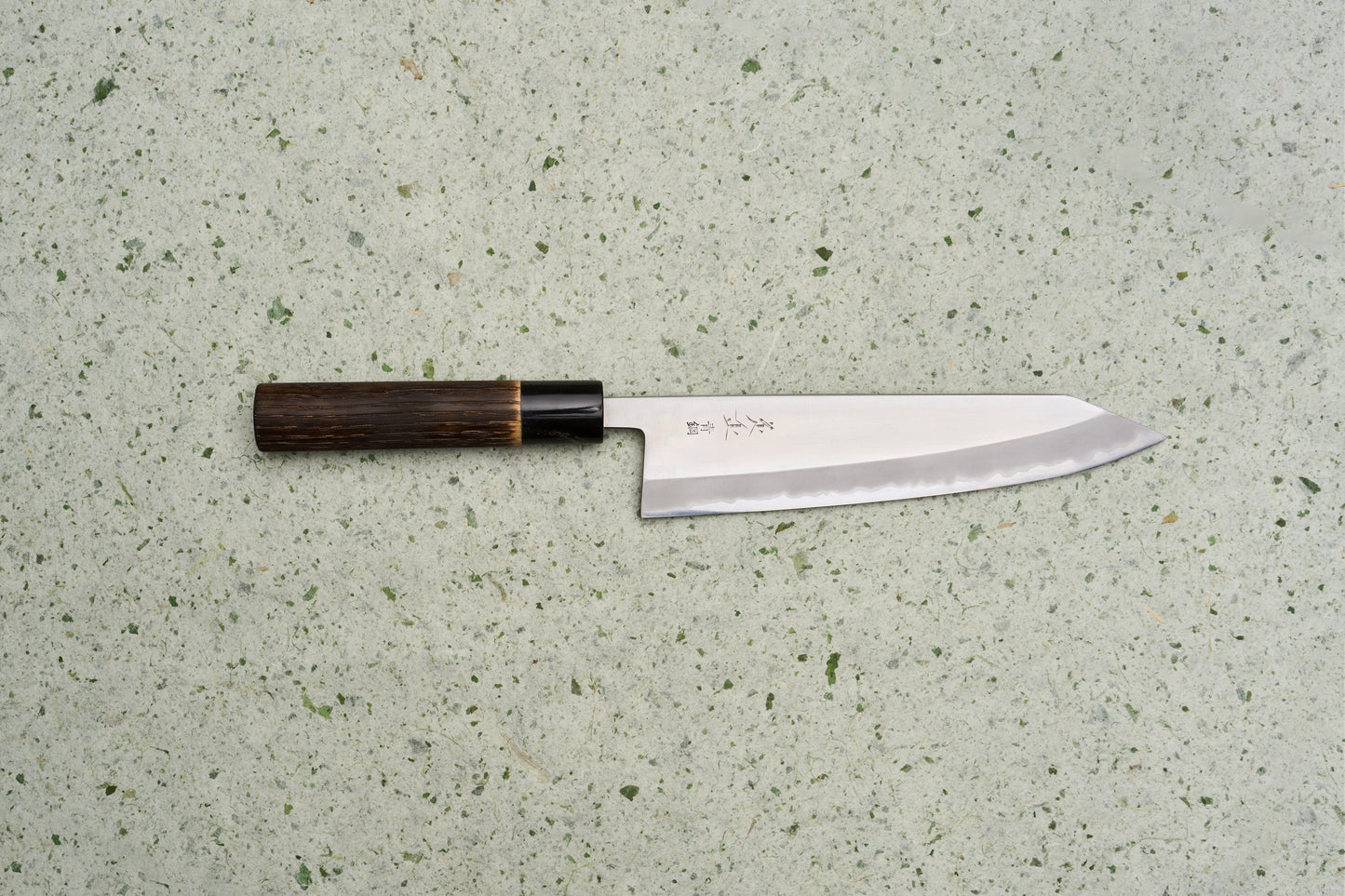 Sadamasa Aogami Stainless Migaki Kiritsuke Gyuto 210mm lies on a speckled countertop, showcasing its high-polish blade with hamon and engraved kanji, paired with a dark octagonal wooden handle.