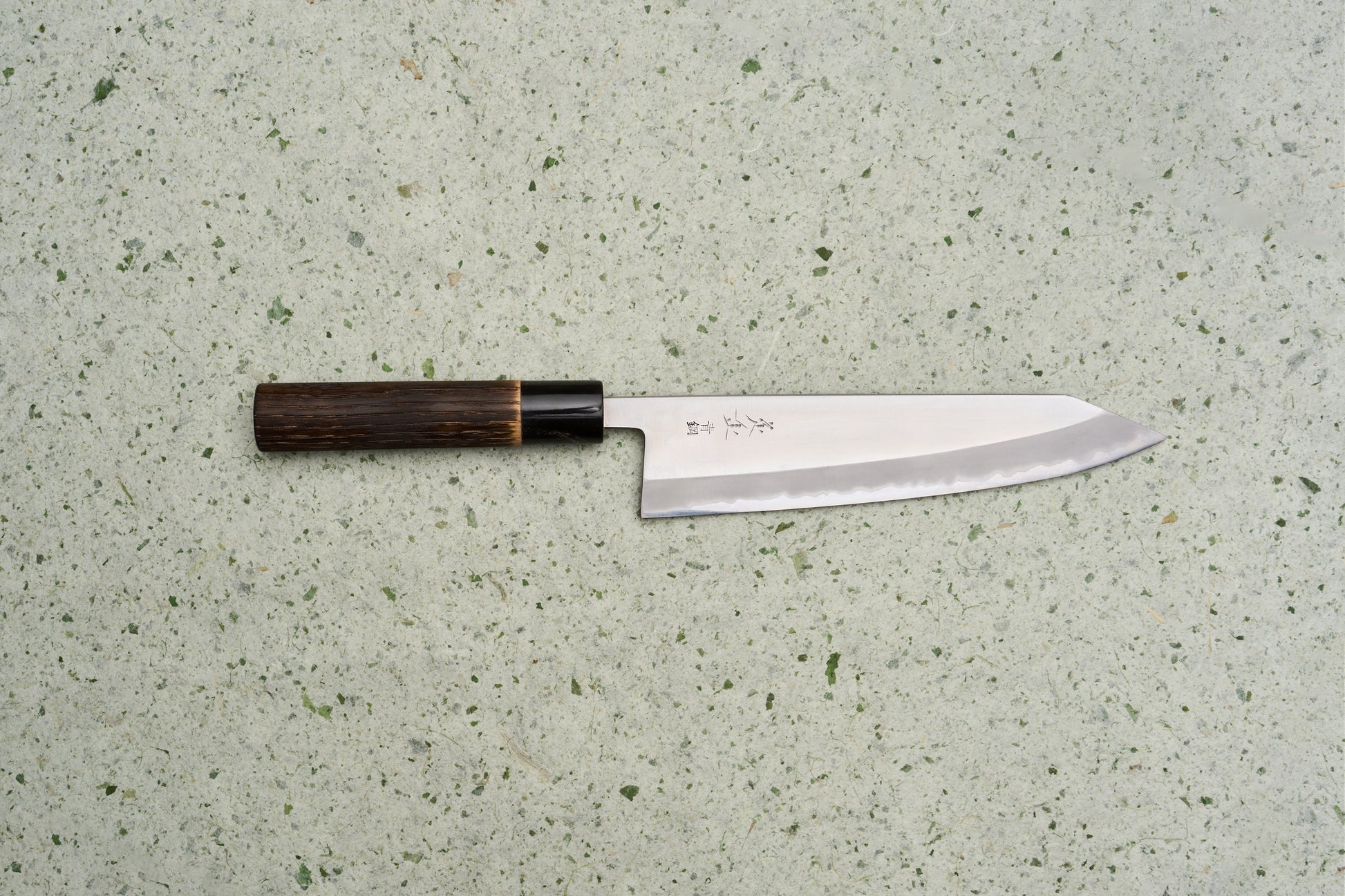 Sadamasa Aogami Stainless Migaki Kiritsuke Gyuto 210mm lies on a speckled countertop, showcasing its high-polish blade with hamon and engraved kanji, paired with a dark octagonal wooden handle.