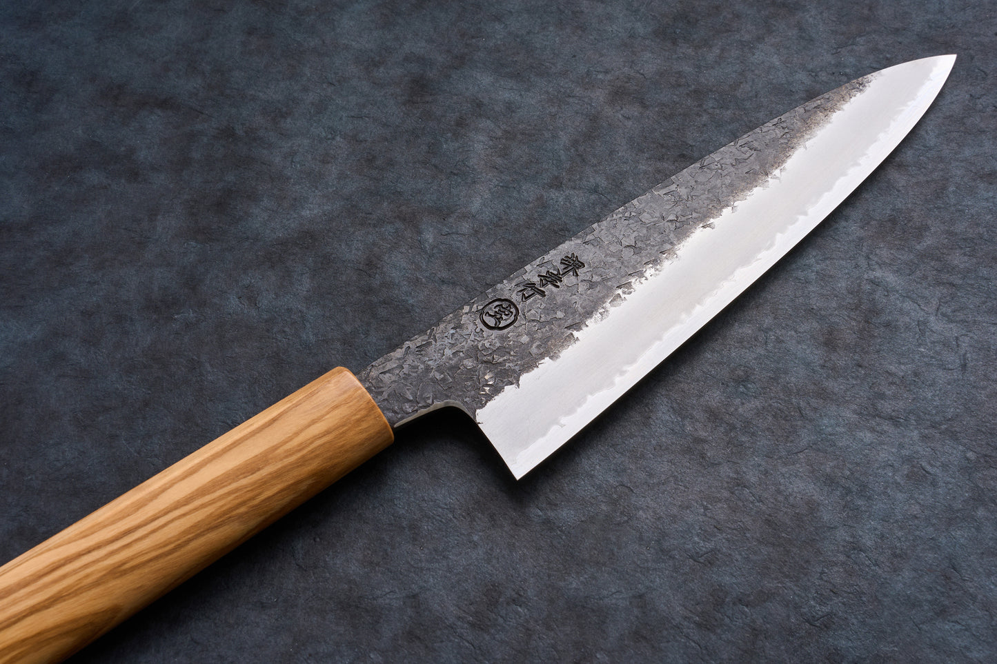 Close-up of Sakai Takayuki Sanpo Mizu Gyuto 210mm, featuring a hammered blade with hamon line and Japanese characters, resting on a slate surface.