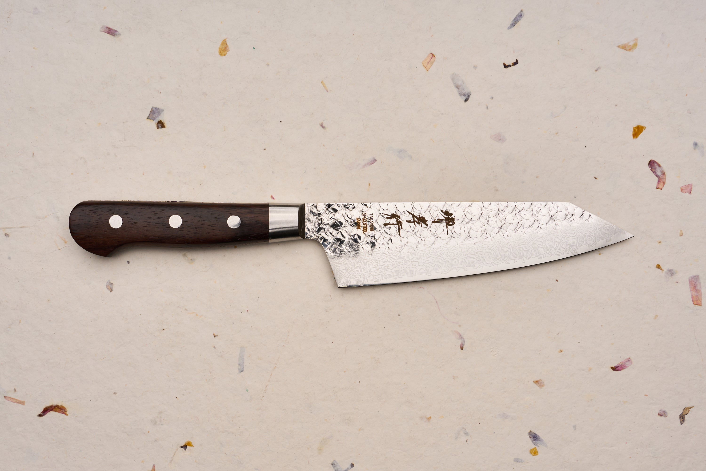 Sakai Takayuki SG2 Tsuchime Damascus Western Kengata Santoku 160mm knife with a dark wooden handle, distinctive hammered blade finish, and engraved Japanese characters, displayed on a textured handmade-paper background.