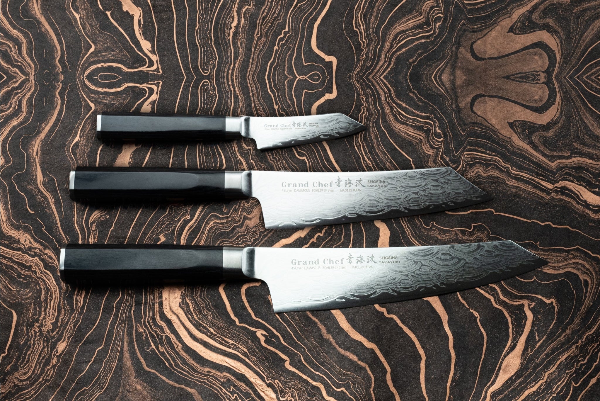 Sakai Takayuki Grand Chef Seigaiha 3 Piece Knife Set on wood-grain surface, featuring octagonal handles and damascus-style blades with Grand Chef stamp; sizes range from utility to chef’s knives.