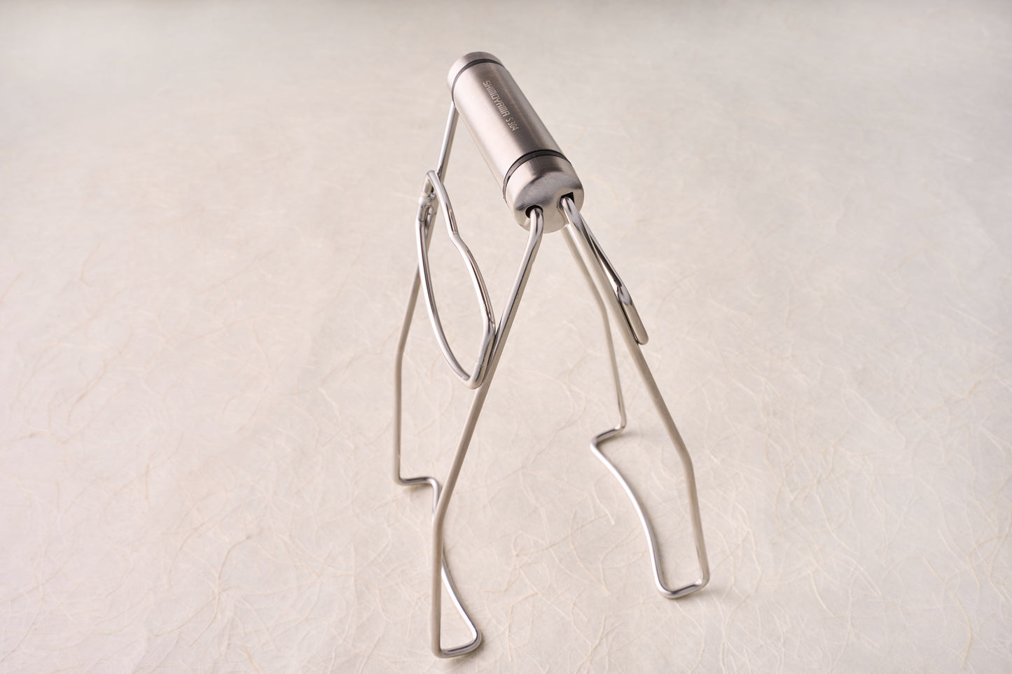 Shimoyama Stainless Steel Dish Clip Lifter, a compact, collapsible stainless-steel tripod ideal for supporting camping cookware or small pans, reflecting Knifewear's commitment to quality chef tools.