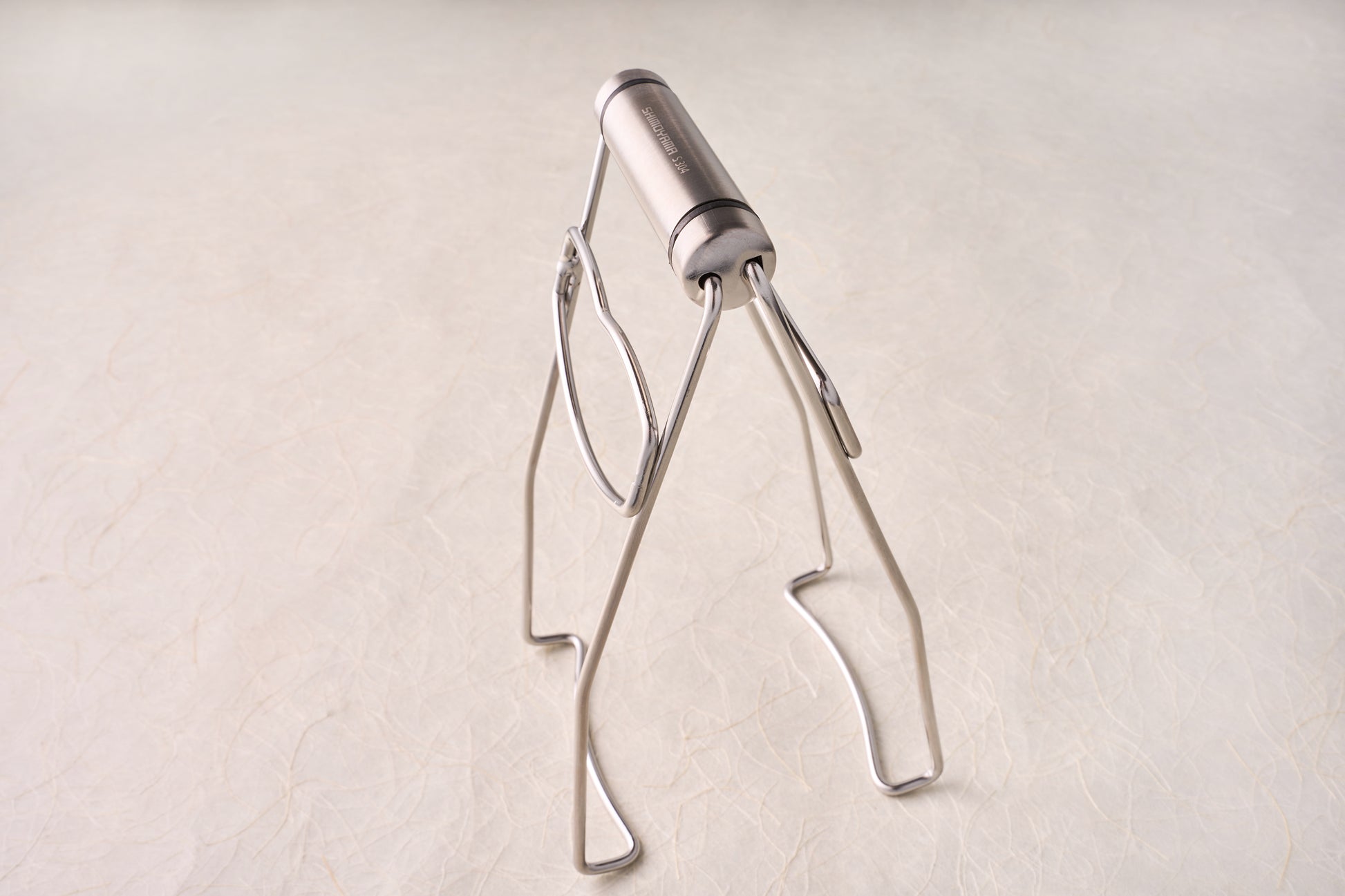 Shimoyama Stainless Steel Dish Clip Lifter, a compact, collapsible stainless-steel tripod ideal for supporting camping cookware or small pans, reflecting Knifewear's commitment to quality chef tools.