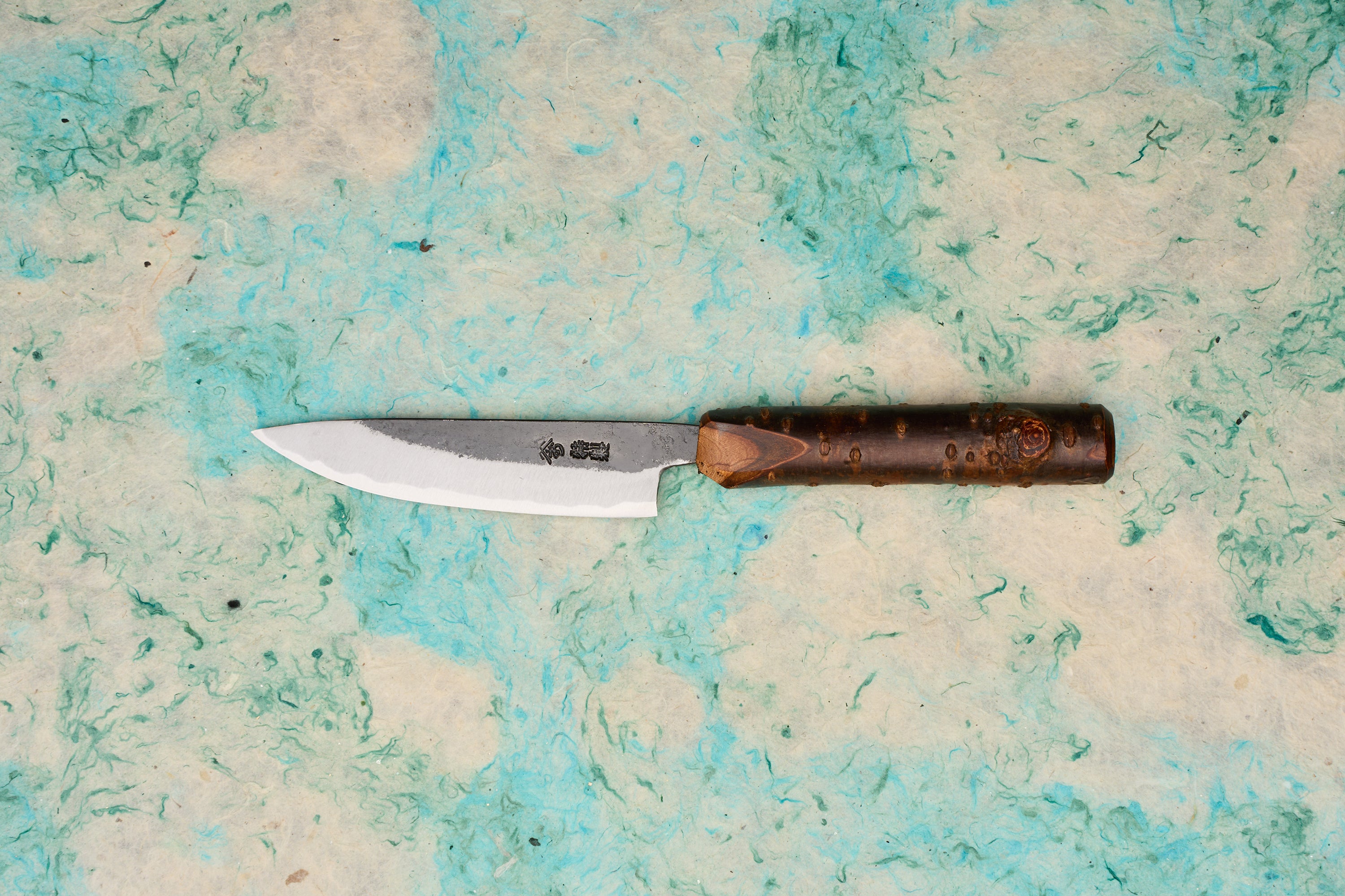 Tottori Otsuka Hamono Aoichi Kurouchi Petty 100mm knife with a single-bevel Aogami #1 steel blade, visible hammer marks, Japanese etchings, and a cylindrical bark-textured wooden handle.