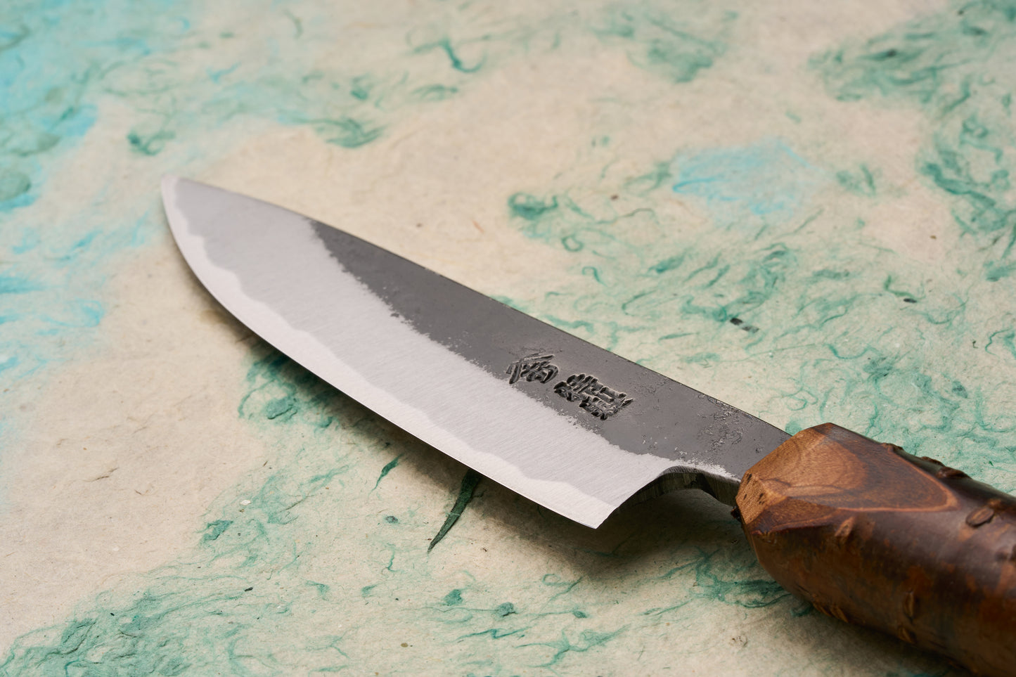 Tottori Otsuka Hamono Aoichi Kurouchi Petty 100mm knife displayed on handmade paper, highlighting its polished cutting edge, rough-forged upper face with Japanese characters, and burl-wood handle, crafted by master blacksmith Otsuka-san.
