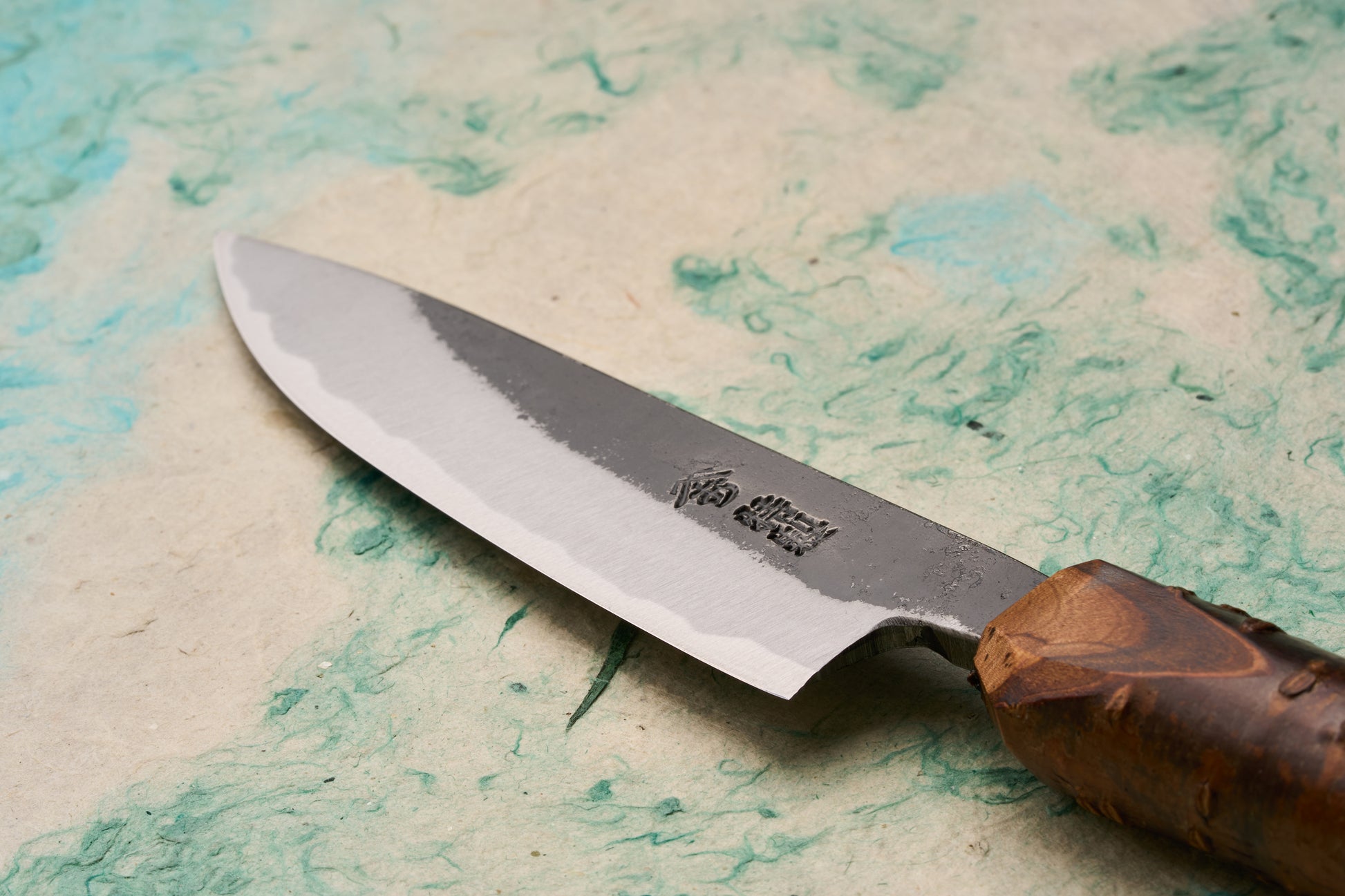 Tottori Otsuka Hamono Aoichi Kurouchi Petty 100mm knife displayed on handmade paper, highlighting its polished cutting edge, rough-forged upper face with Japanese characters, and burl-wood handle, crafted by master blacksmith Otsuka-san.
