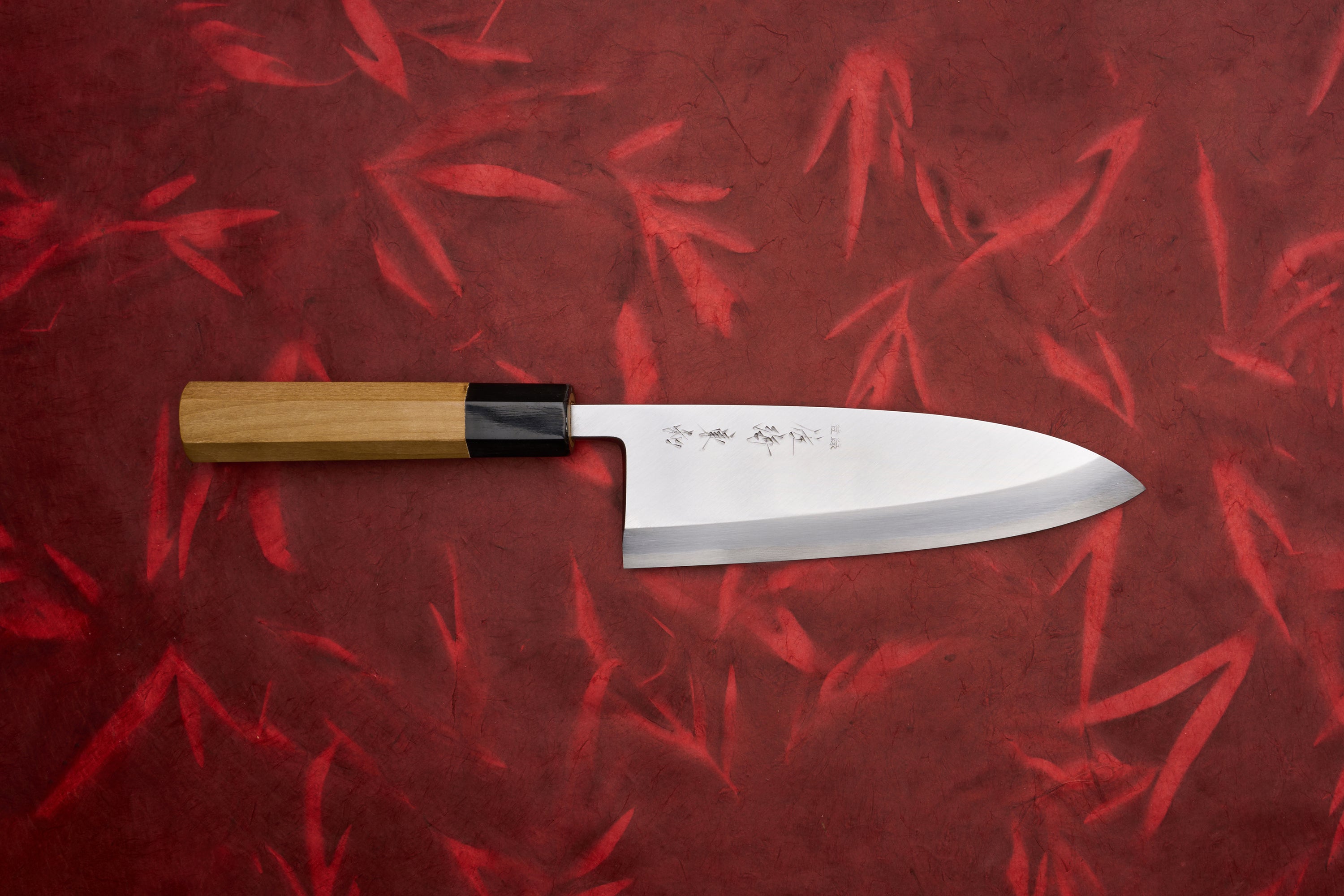 Touroku Sakai Tohwa Shirogami Deba 180mm, a Japanese kitchen knife with an engraved stainless steel blade and octagonal wooden handle, showcased on a red textured background.