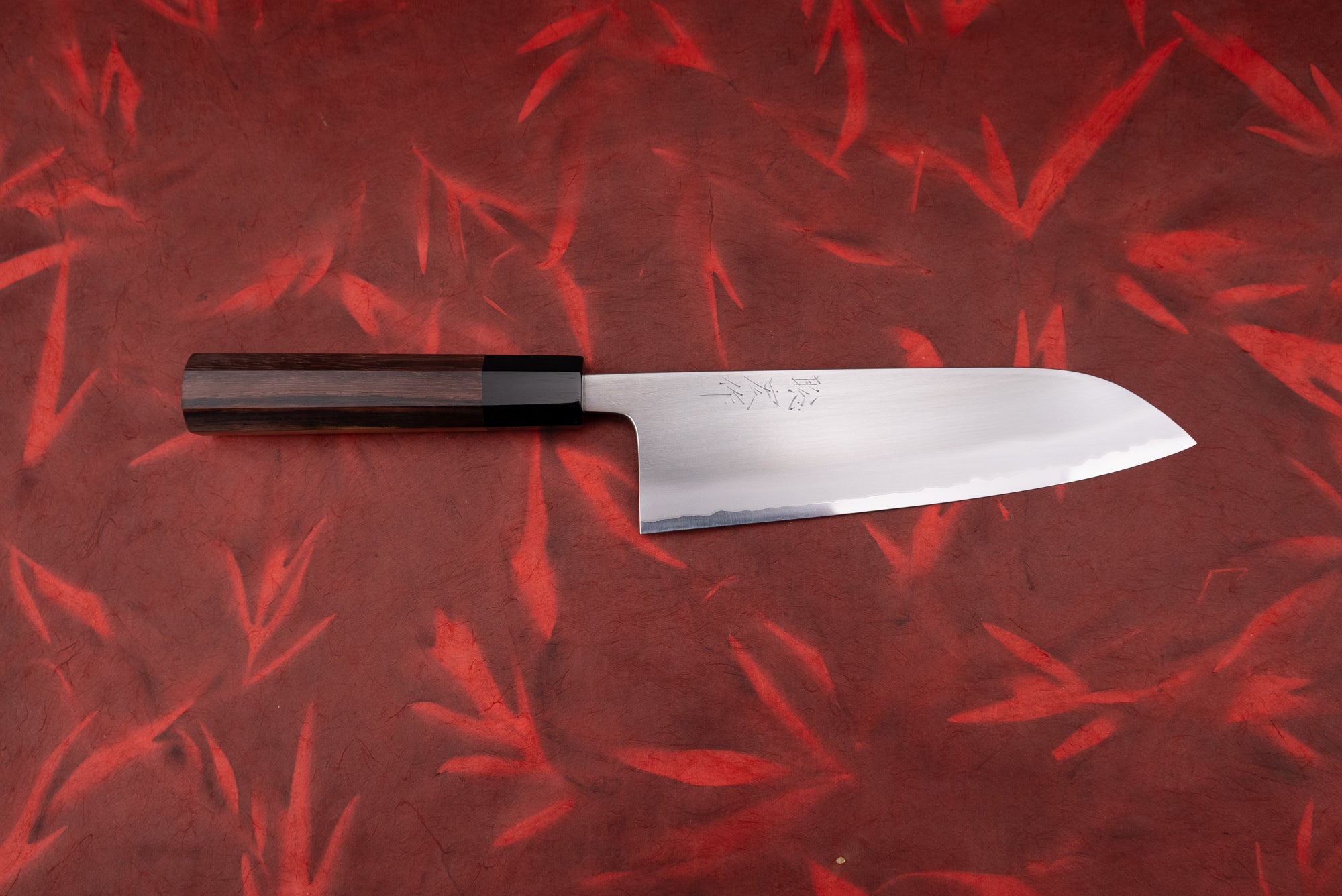 Toshihiro Wakui Shirogami Stainless Migaki Santoku 180mm, featuring a straight-edged stainless steel blade and dark wood handle, displayed on a red patterned surface. A multi-purpose Japanese kitchen knife.