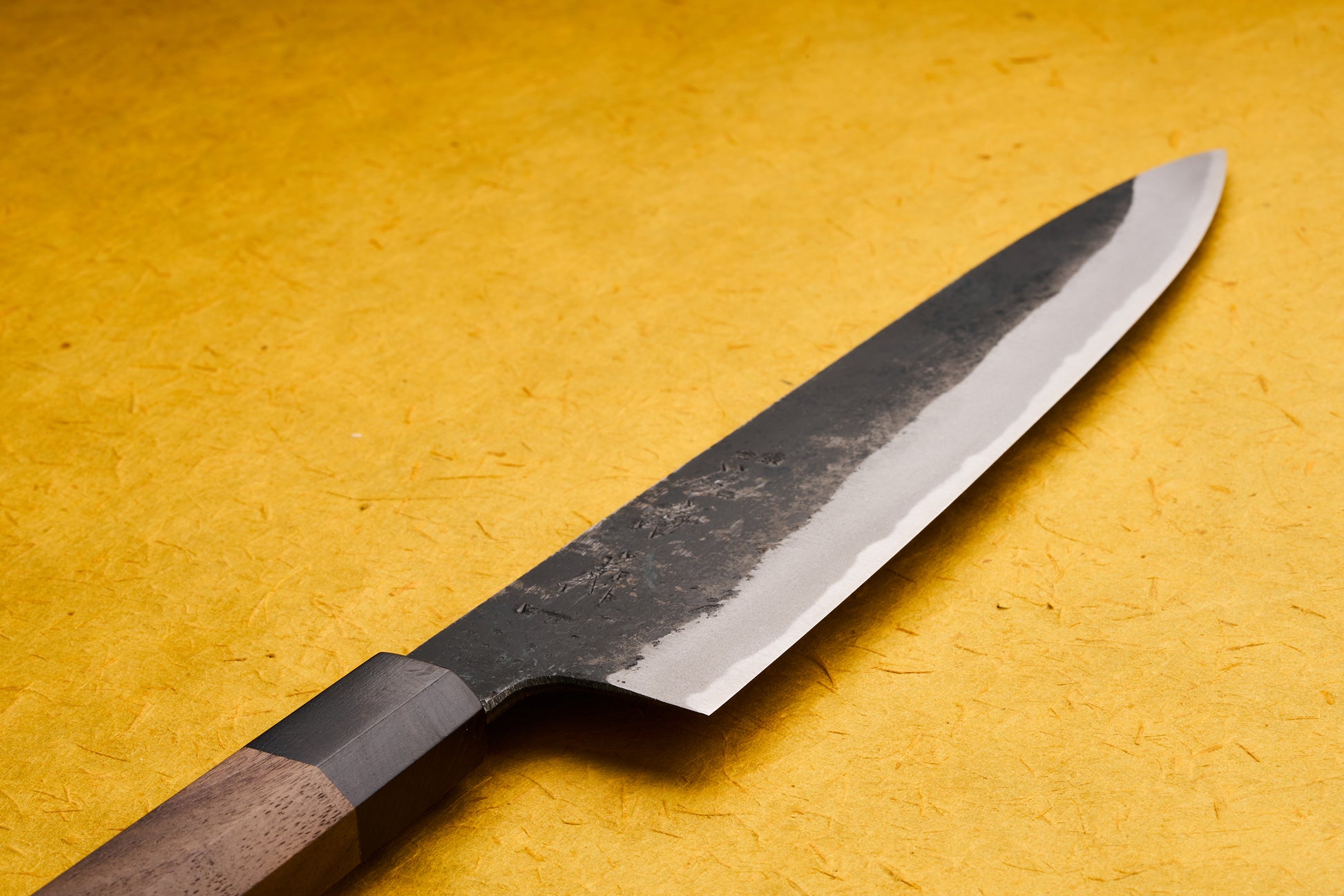 Touroku Sakai Yoshikazu Shirogami Kurouchi Gyuto 240mm knife with a high-carbon blade featuring a rough-forged patina and a polished edge, laid on a textured yellow mat, showcasing its elegant wooden handle.