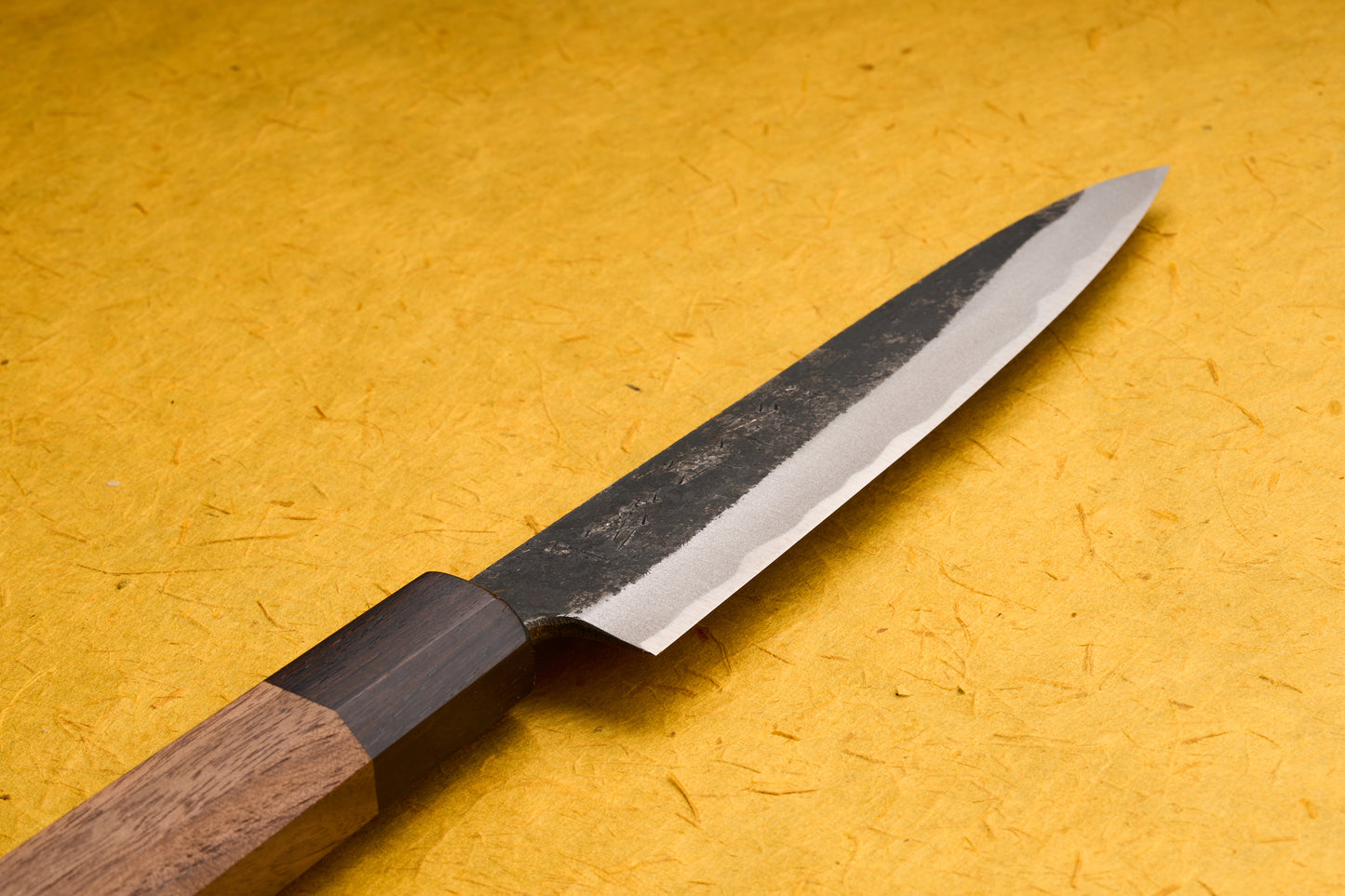 Touroku Sakai Yoshikazu Shirogami Kurouchi Petty 150mm, a Japanese-style kitchen knife, features a dark, hammered blade and a polished edge with a two-tone octagonal wood handle, ideal for precise cutting.