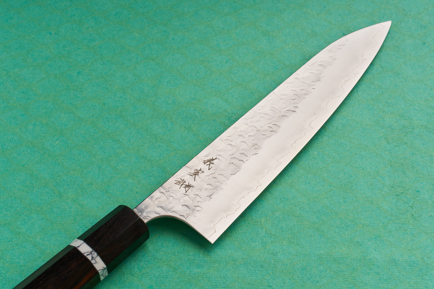 Yoshimi Kato SG2 Hyo Gyuto 210mm featuring a hammered damascus finish, subtle hamon line, and octagonal wooden handle with a decorative ferrule, showcased on a teal background.