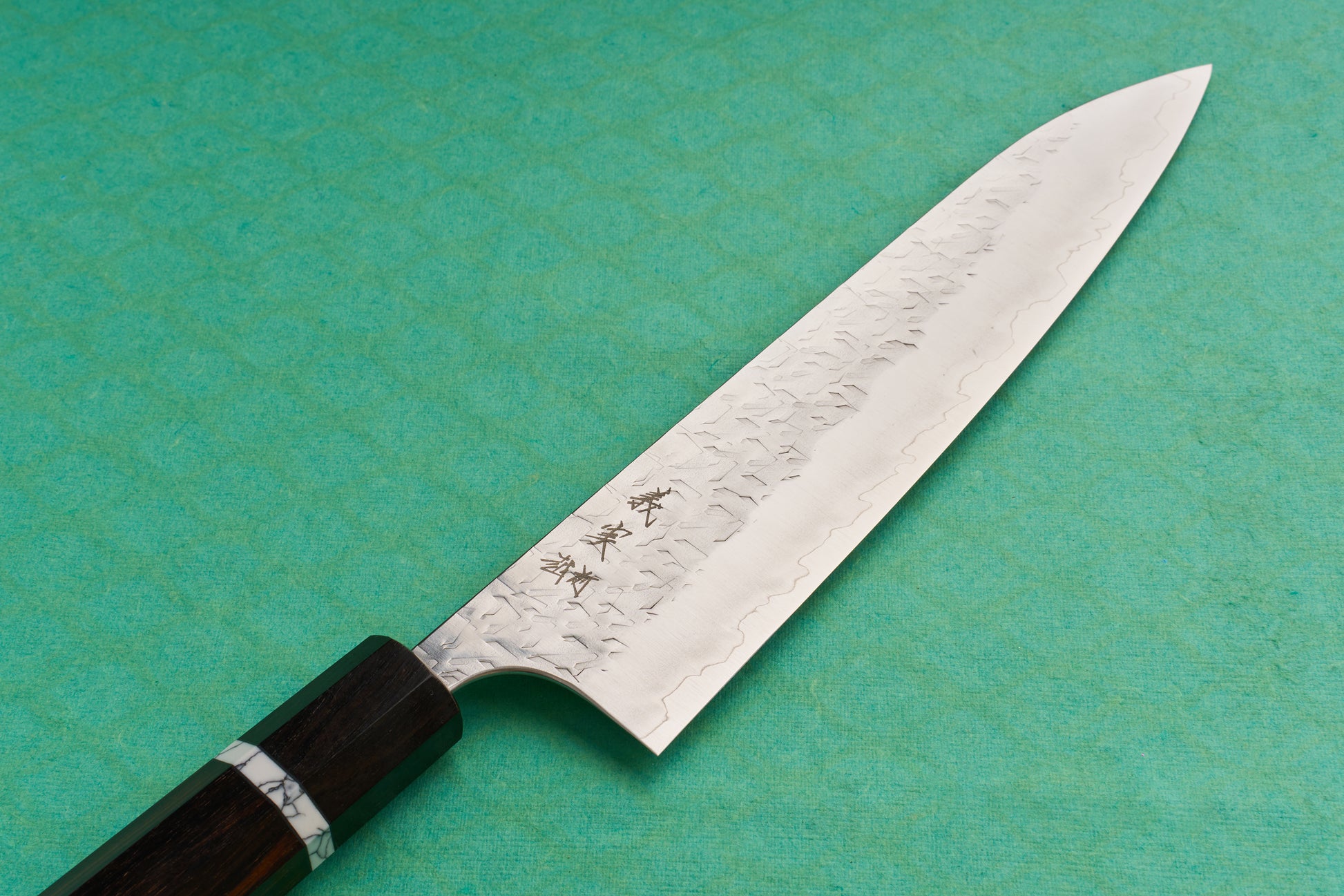 Yoshimi Kato SG2 Hyo Gyuto 210mm featuring a hammered damascus finish, subtle hamon line, and octagonal wooden handle with a decorative ferrule, showcased on a teal background.