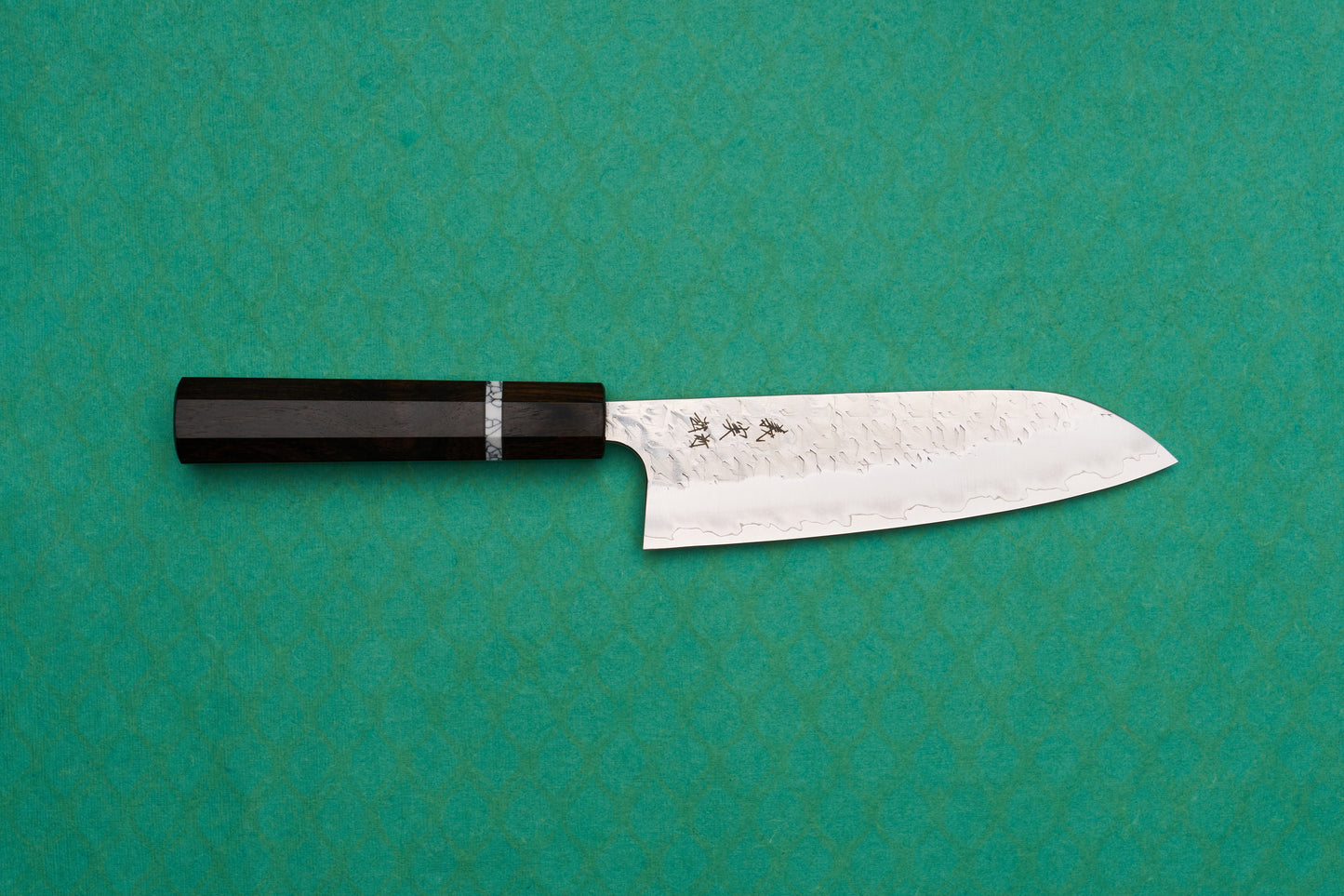 Yoshimi Kato SG2 Hyo Santoku 165mm knife with a hammered finish and kanji etching, featuring a dark wood octagonal handle, displayed on a green, patterned backdrop.
