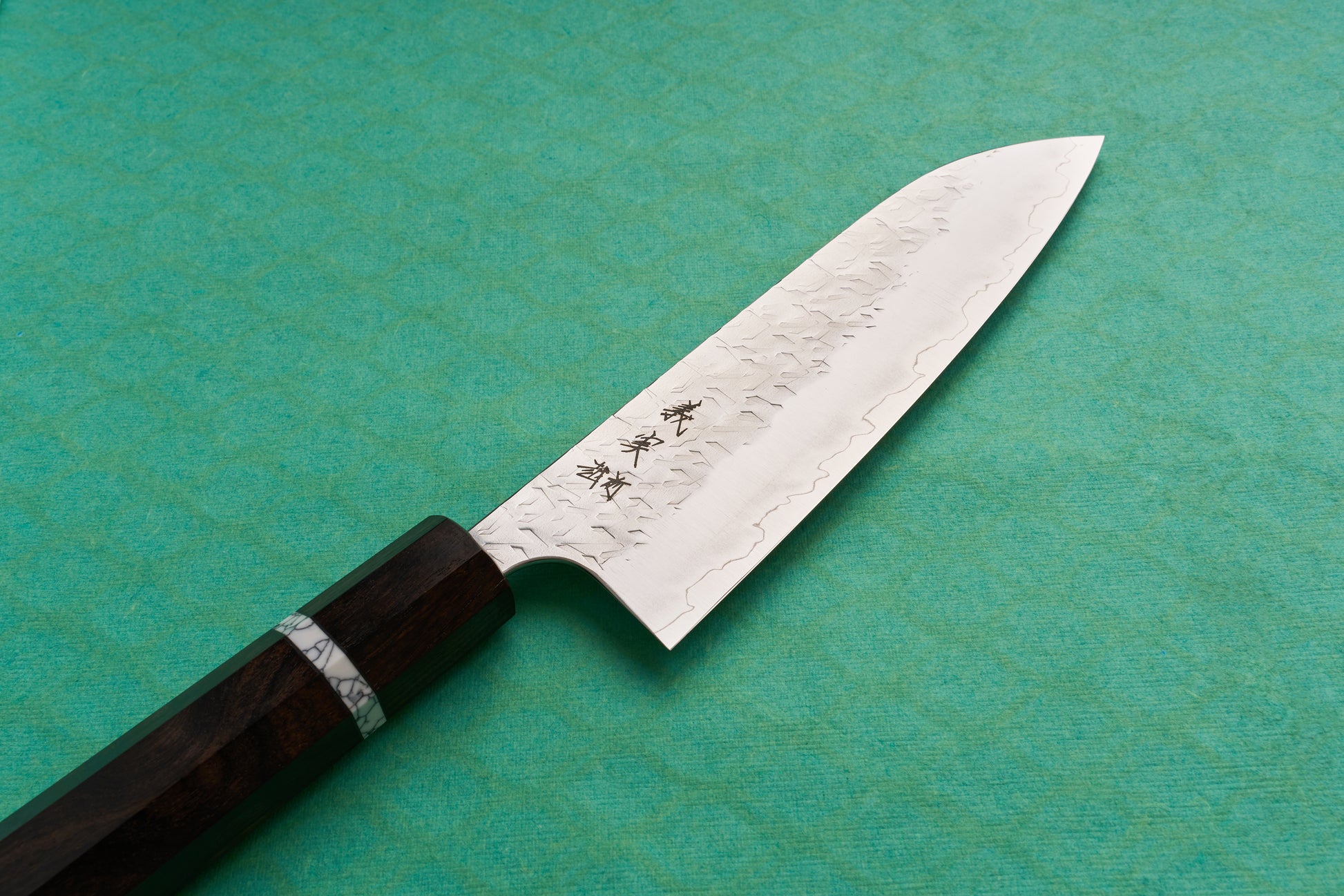 Yoshimi Kato SG2 Hyo Santoku 165mm knife with Damascus patterns, hammer marks, engraved Japanese characters, and a dark wooden octagonal handle, displayed on a teal-green surface.
