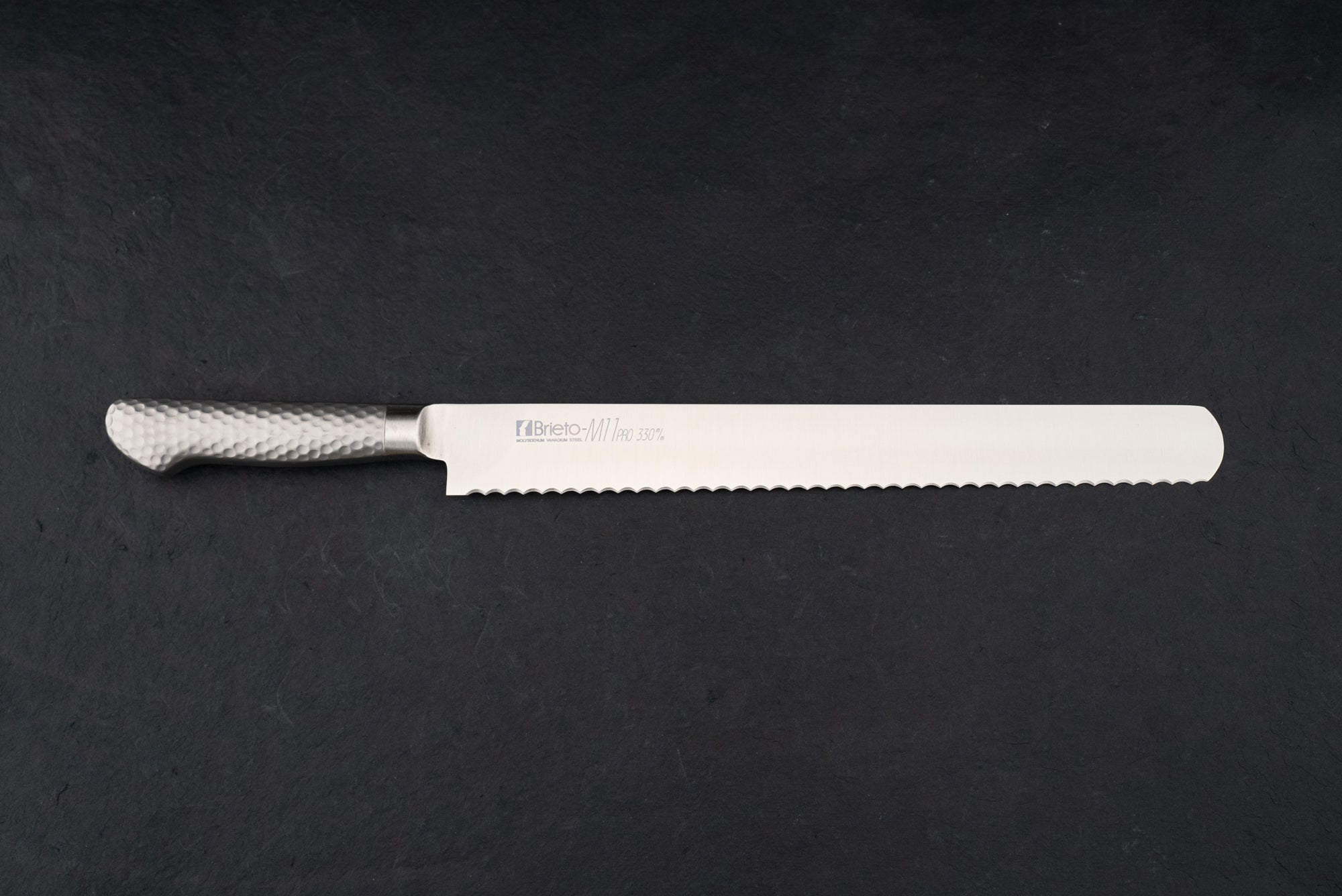 Brieto Pro Bread Knife 330mm M1150, featuring a long, serrated stainless-steel blade and a hammered-metal handle, displayed flat on a dark surface, epitomizing the precision and craftsmanship of Japanese kitchen knives.
